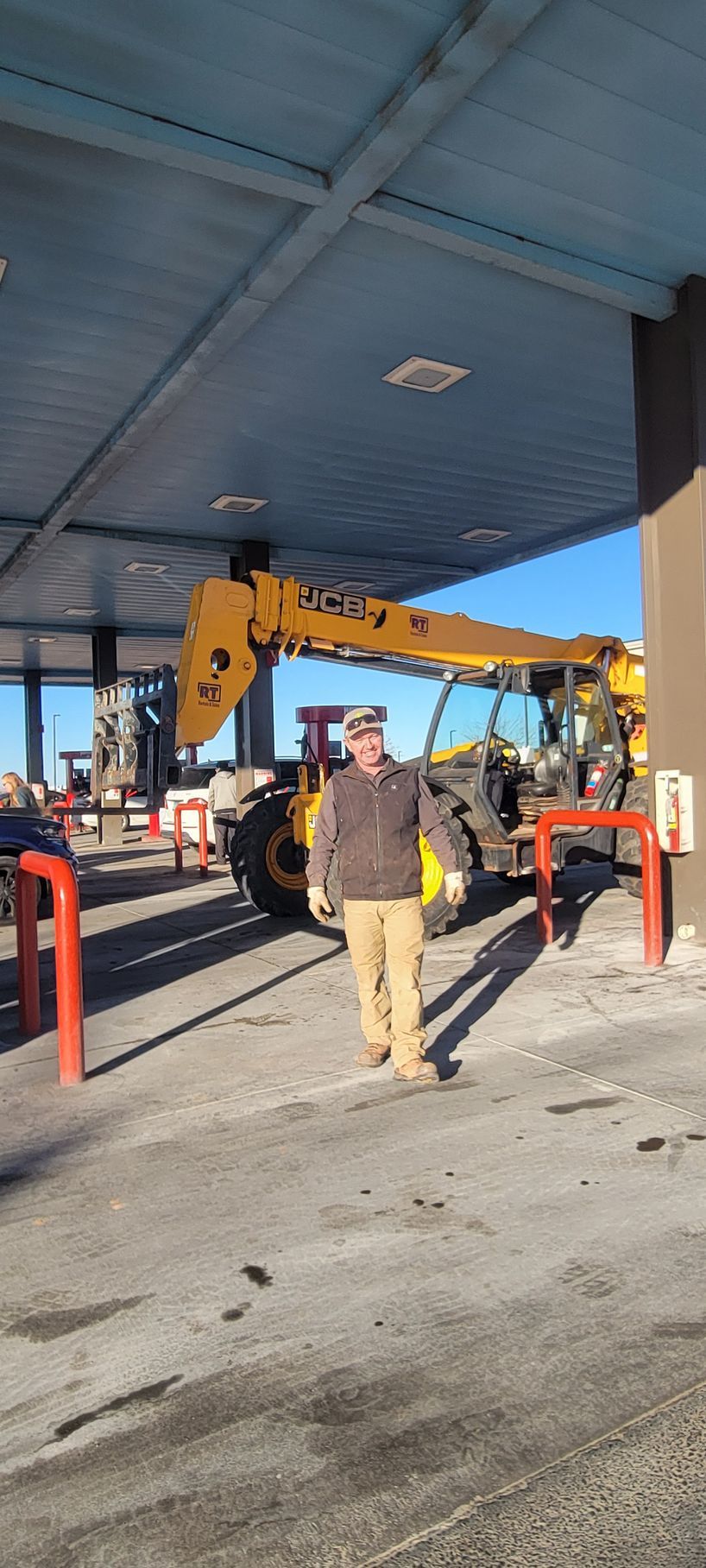 A person stands smiling in front of a yellow telescopic forklift under a gas station canopy.