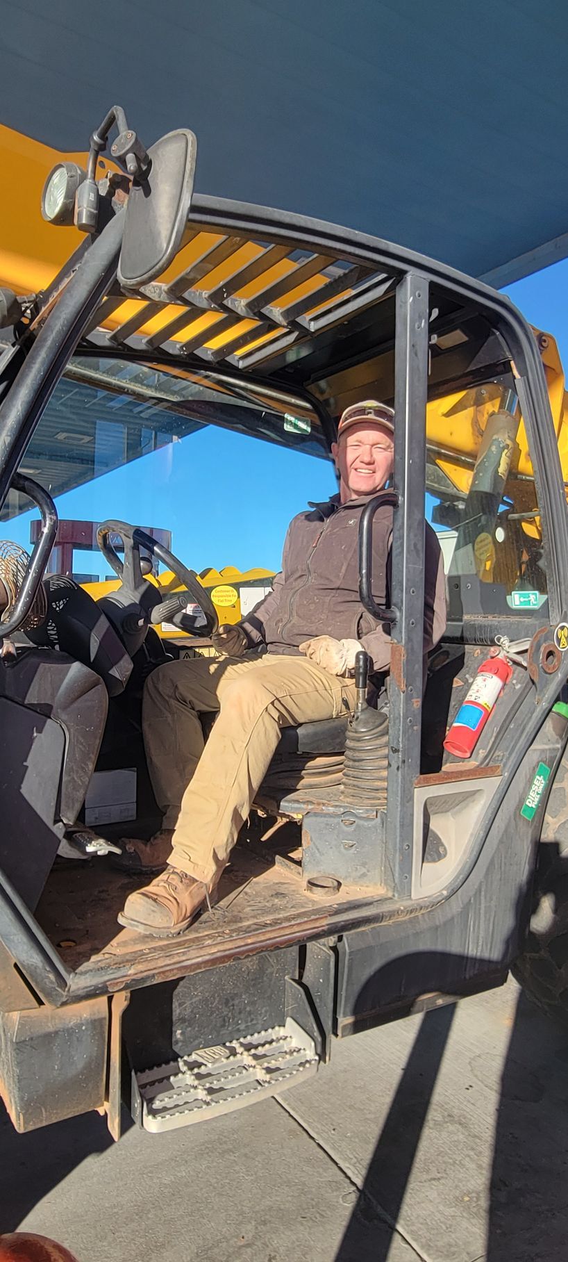 A person wearing a brown jacket, tan pants, and a cap sits in the operator's seat of a yellow construction machine.