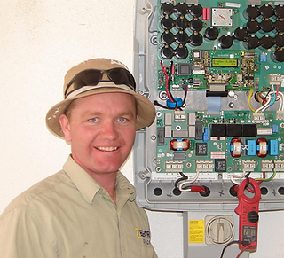 A smiling technician in a hat next to an open electronic control panel with a digital multimeter attached.