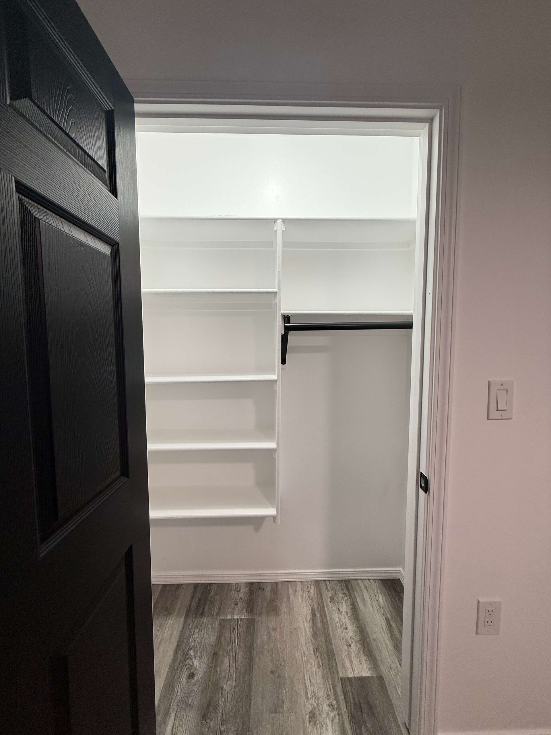 An open black door looks into a walk-in closet with white shelves and a clothing rod against a white back wall.