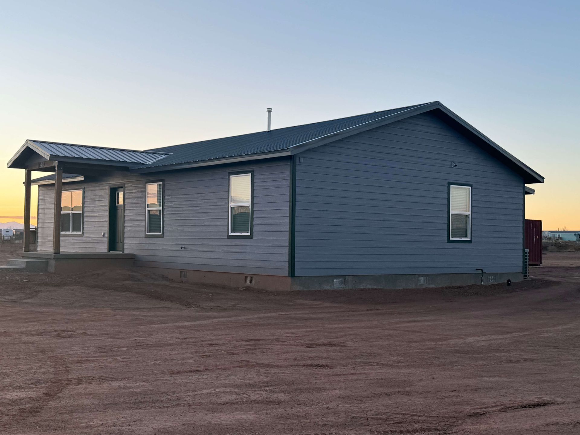 A small, single-story house with a red metal roof, light-colored stucco walls, and a covered front porch on a dirt lot.