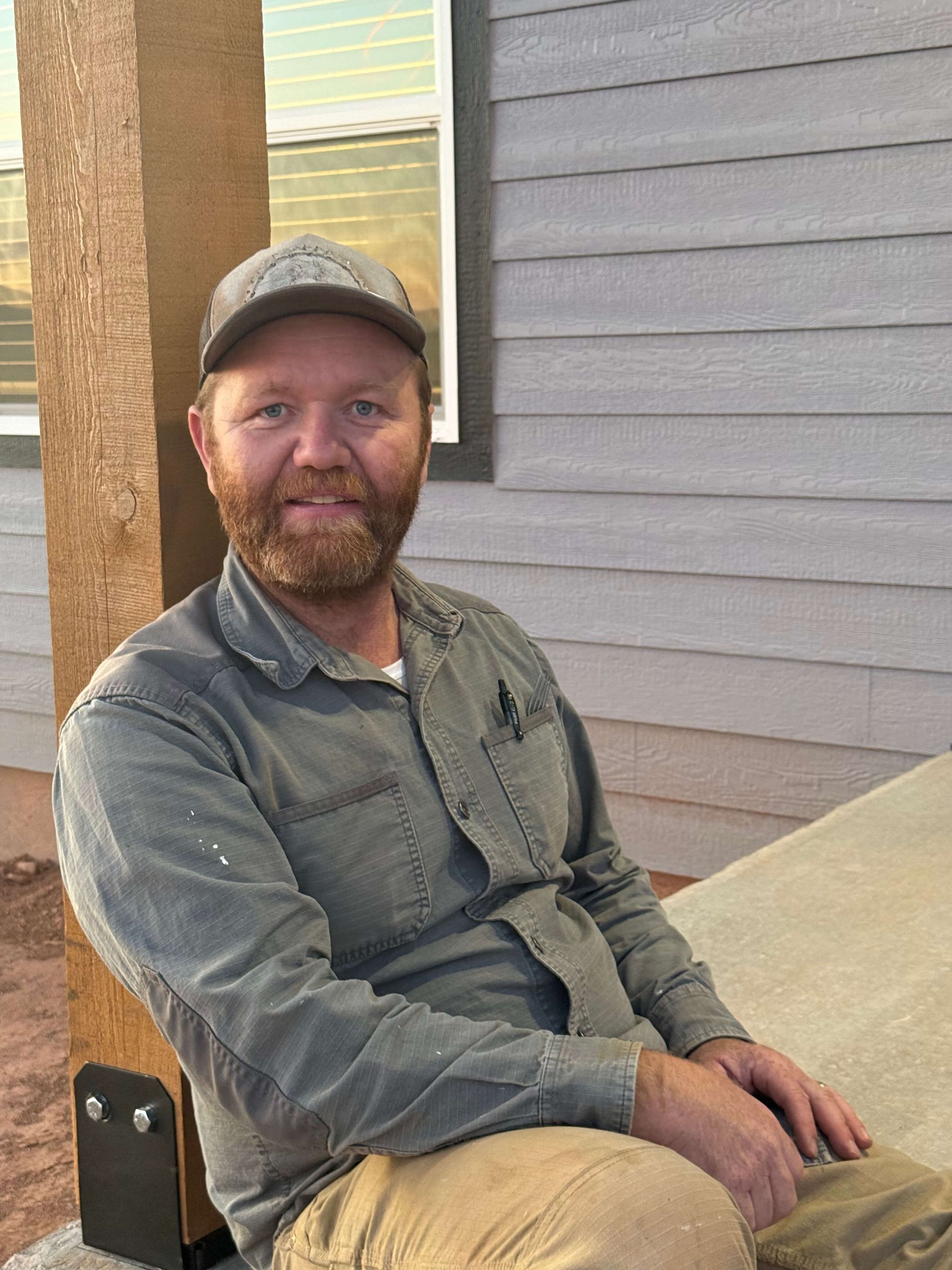 A person wearing a grey shirt and cap sits outside by a wooden porch post and grey siding.