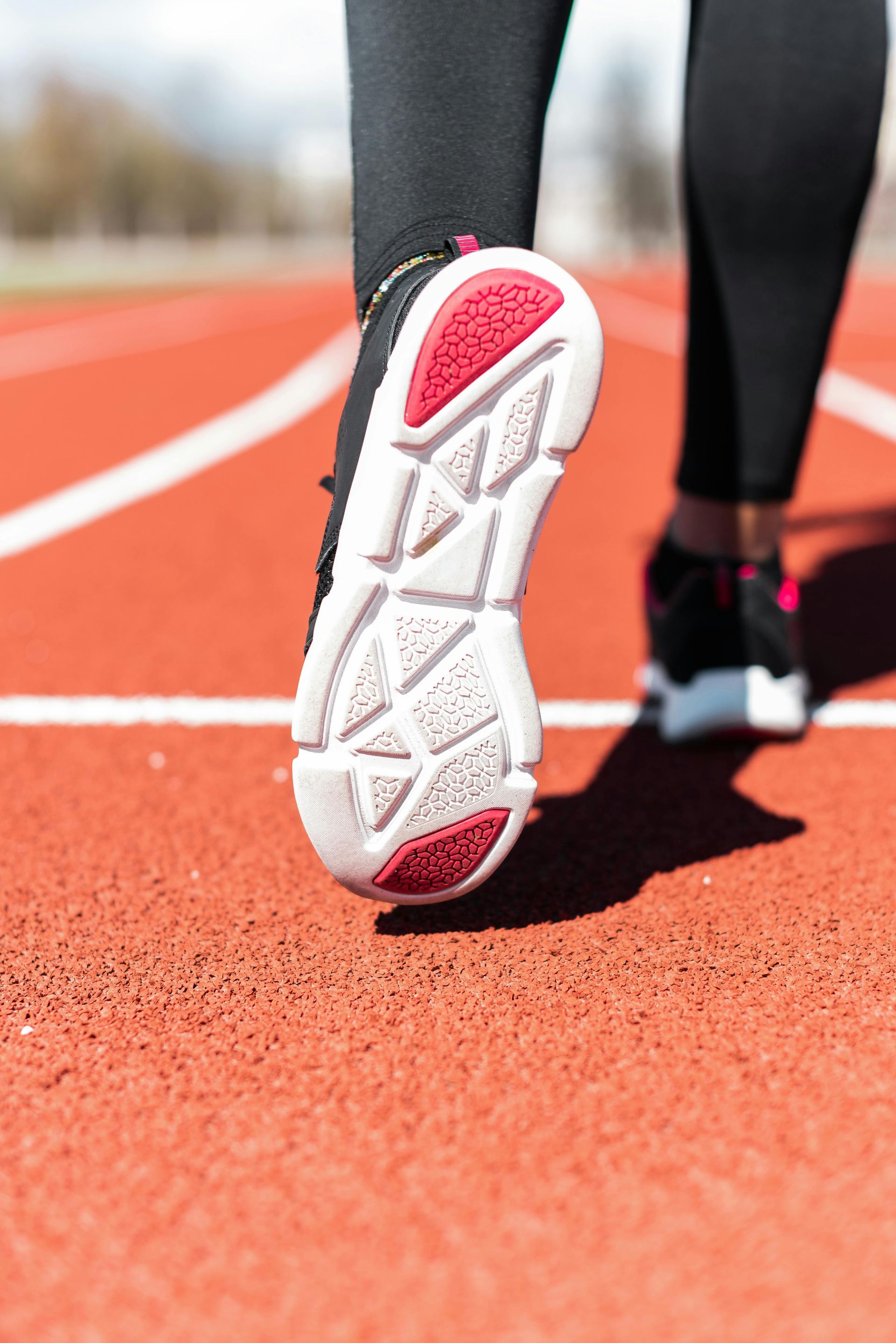 Close-up van een hardloopschoen op een rode atletiekbaan, met een andere hardloper wazig op de achtergrond.