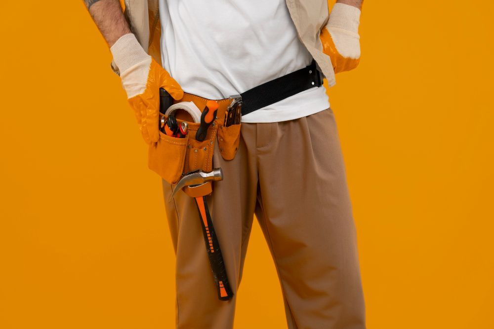 Persona que lleva guantes de trabajo y un cinturón de herramientas, sosteniendo un martillo, sobre un fondo naranja.