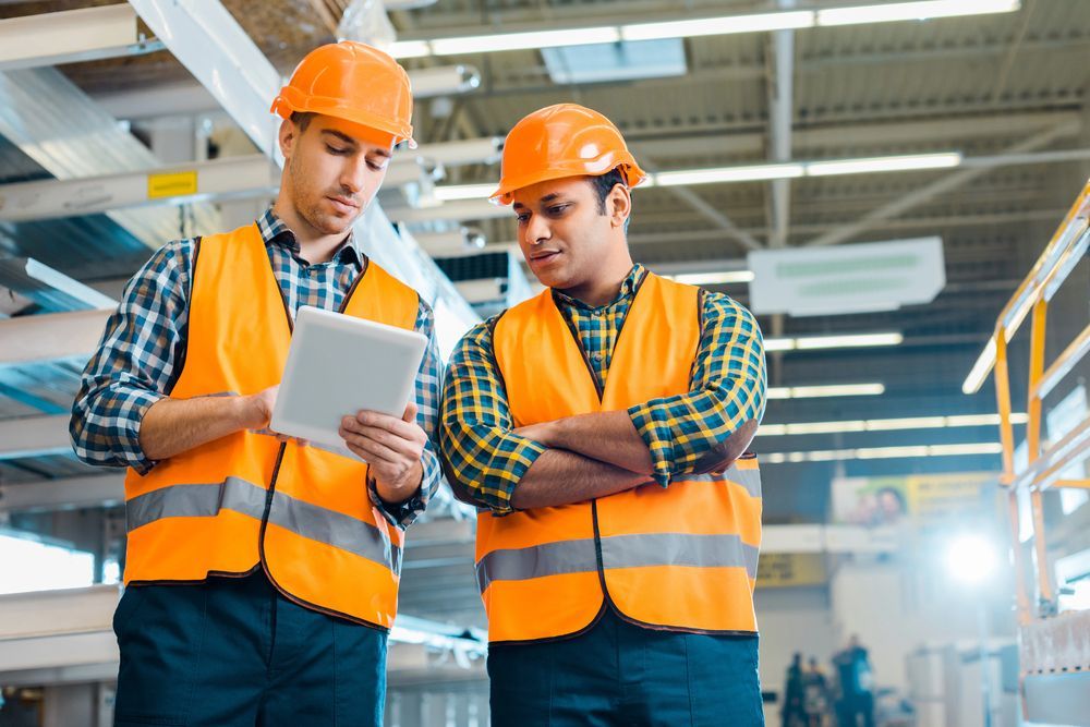 Dos trabajadores de la construcción con chalecos y cascos naranjas miran una tableta en un almacén.