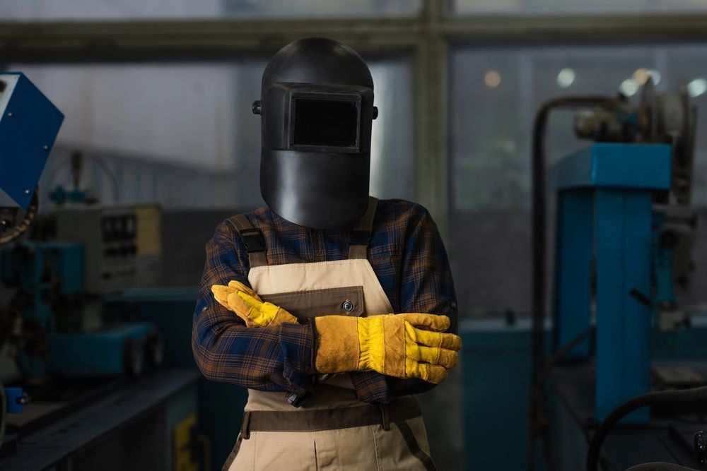 Soldador en un taller con mascarilla, guantes y delantal y con los brazos cruzados.
