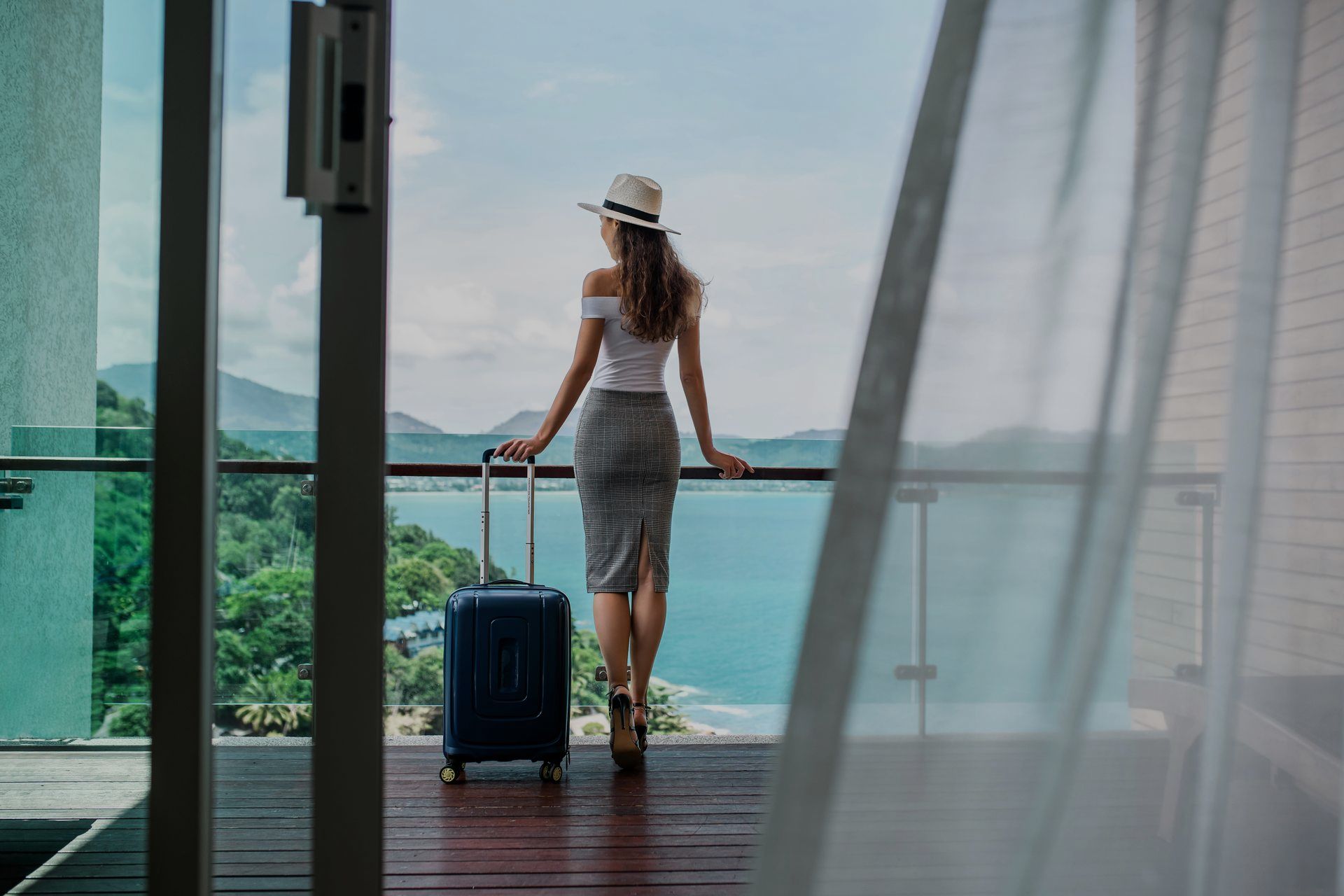 A woman with a suitcase is standing on a balcony overlooking the ocean.