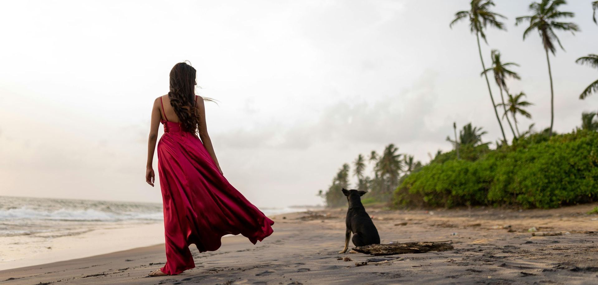 A woman in a red dress is walking on the beach with a black dog.