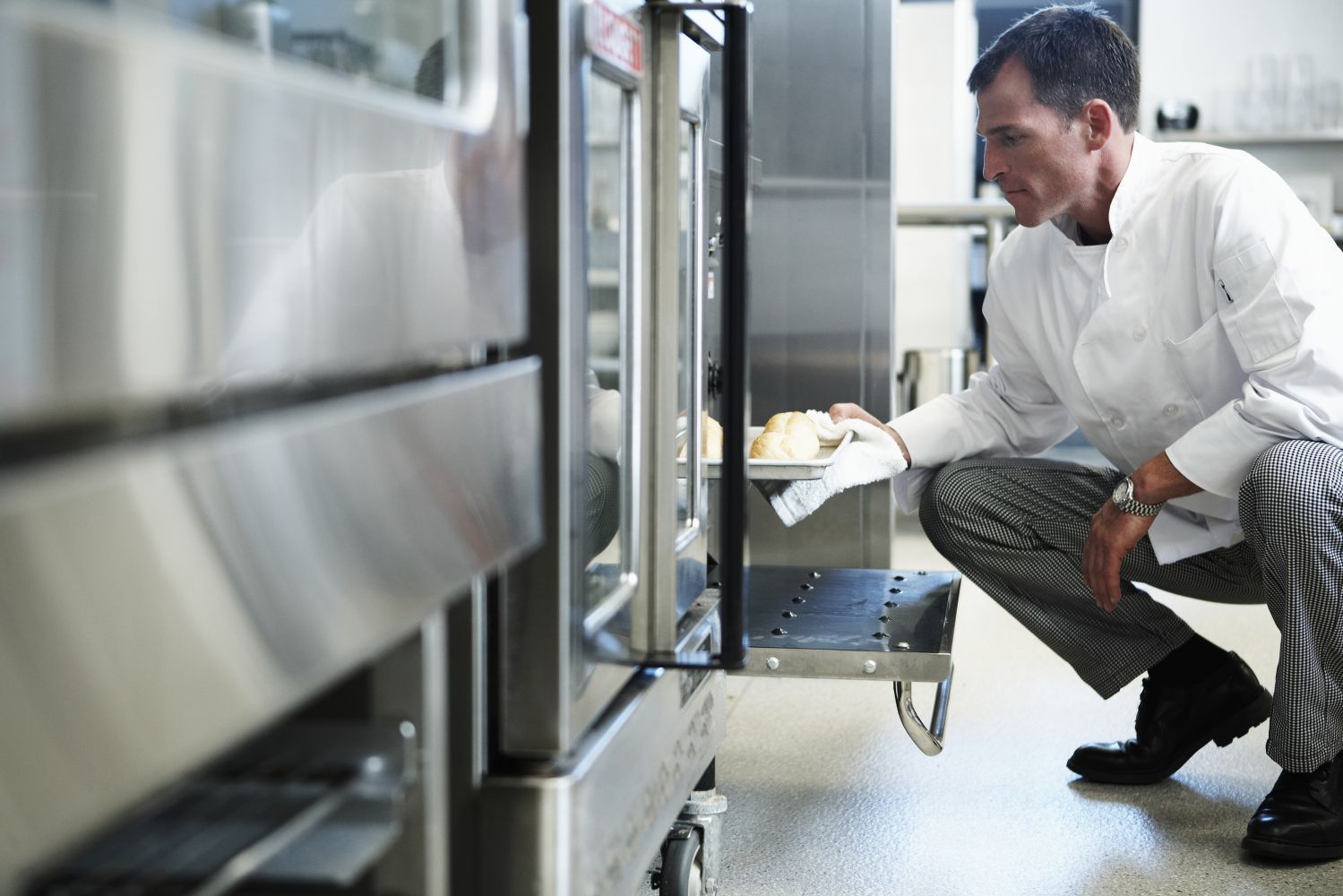 Man in white shirt crouches by an open industrial oven, checking food trays in a commercial kitchen.