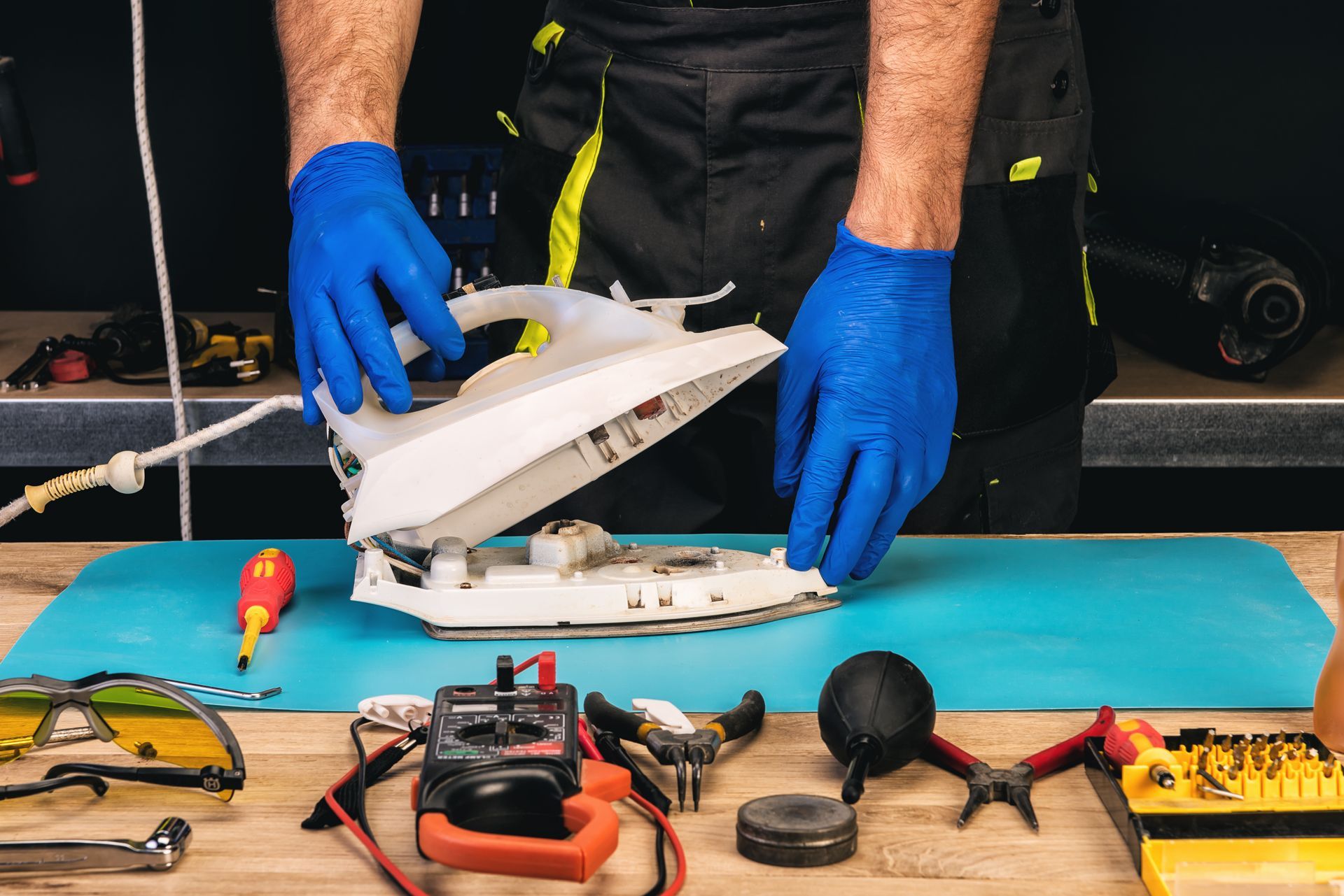 Blue-gloved hands repairing a white appliance at a workbench with scattered tools and parts