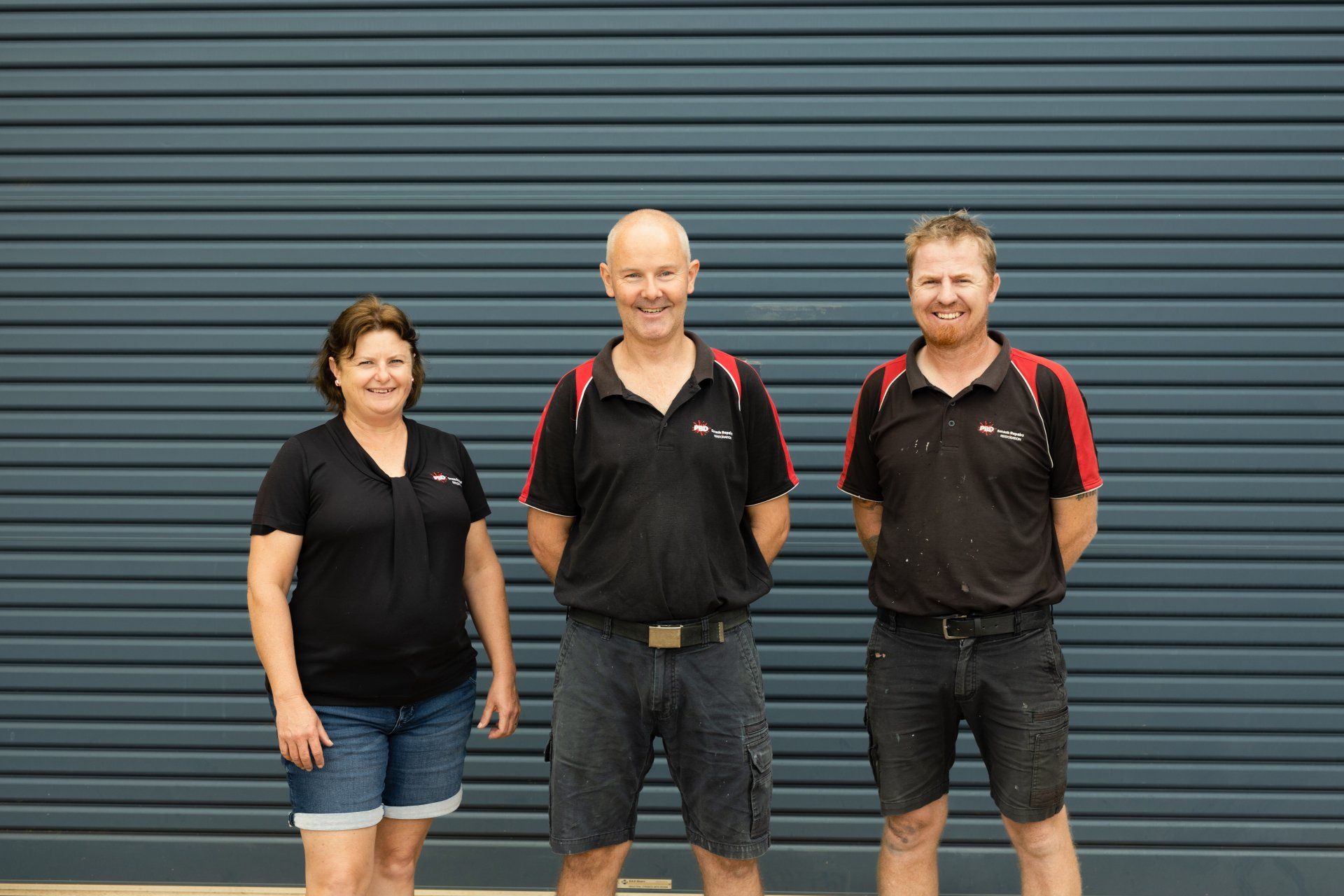 Three people standing in front of a blue corrugated wall, wearing black shirts and shorts.