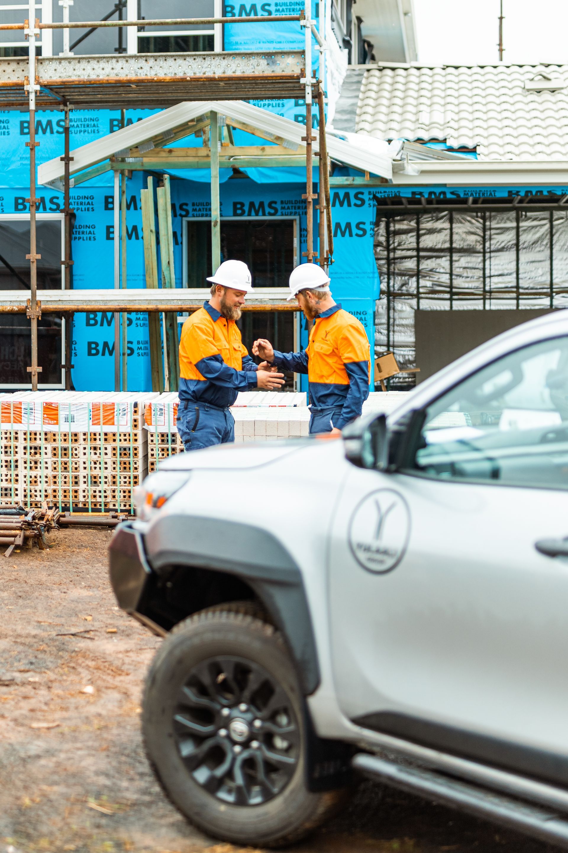 Two construction workers in hard hats stand by a white pickup truck at a building site.