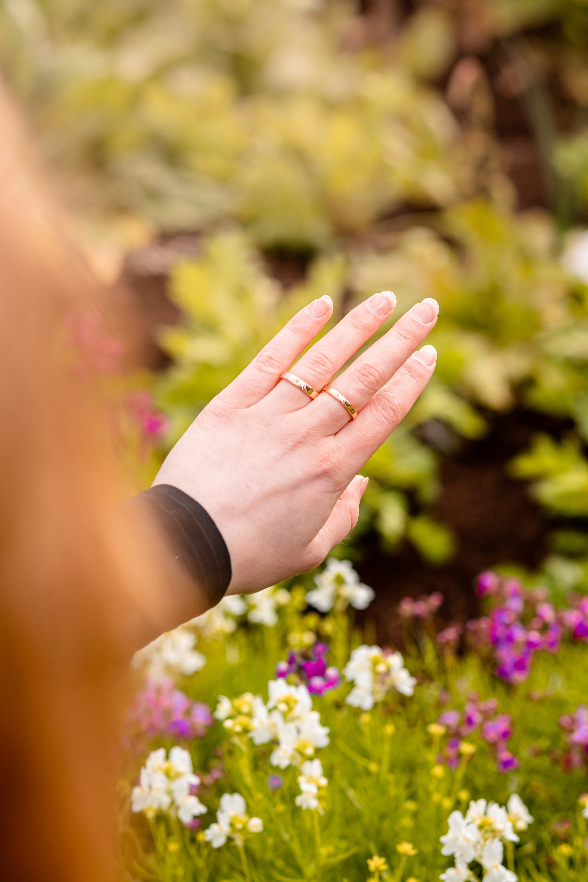 Hand reaching toward small white and purple flowers in a garden with green foliage.