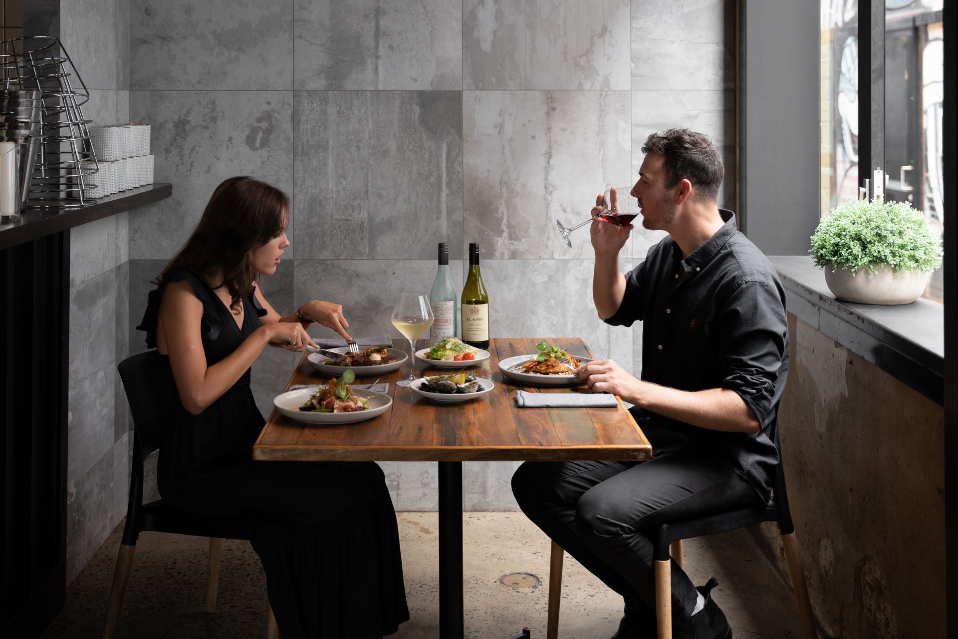 Two people dining at a restaurant table by a window, sharing plates and drinks.