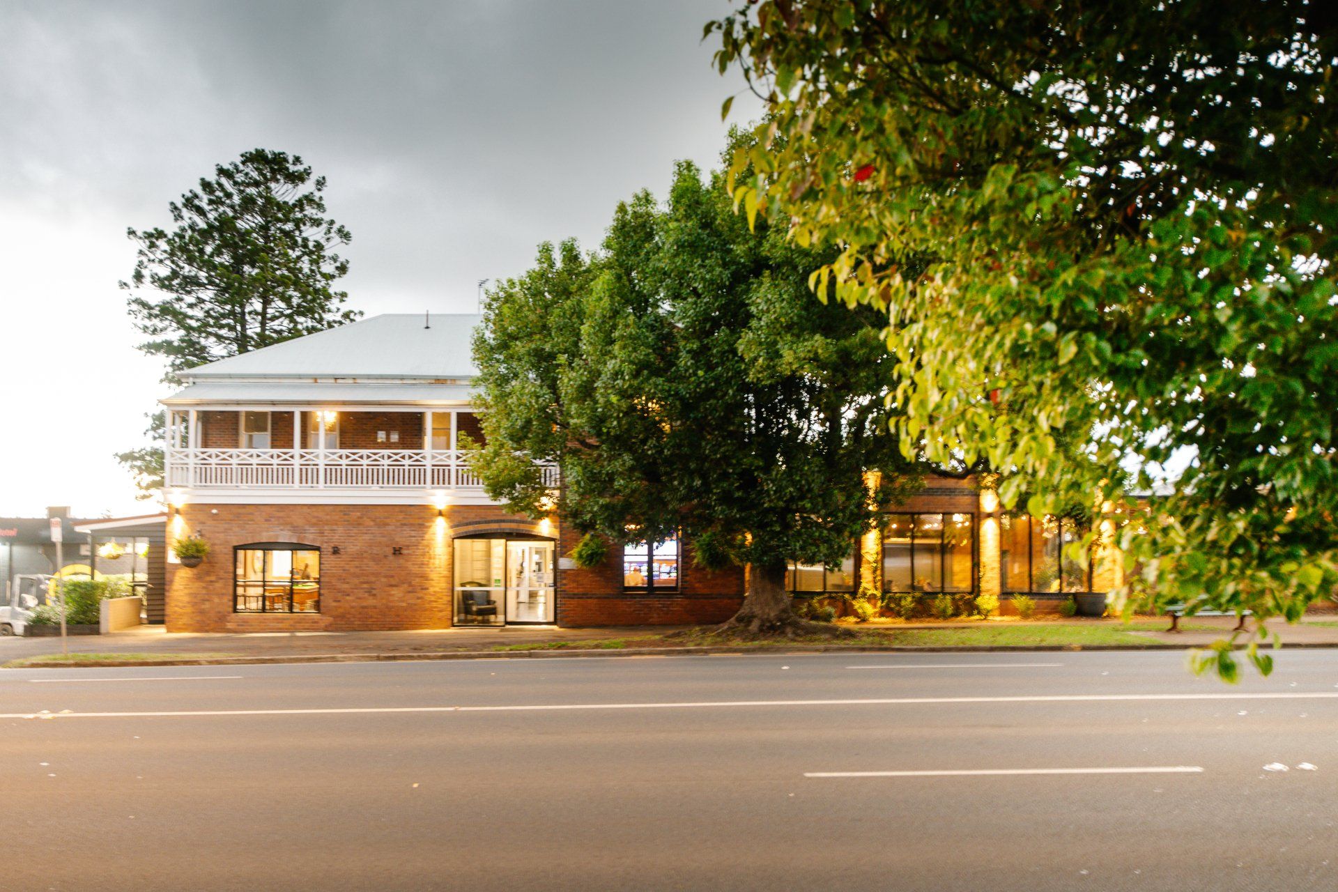 Two-story brick building with lit windows and trees along a quiet street at dusk
