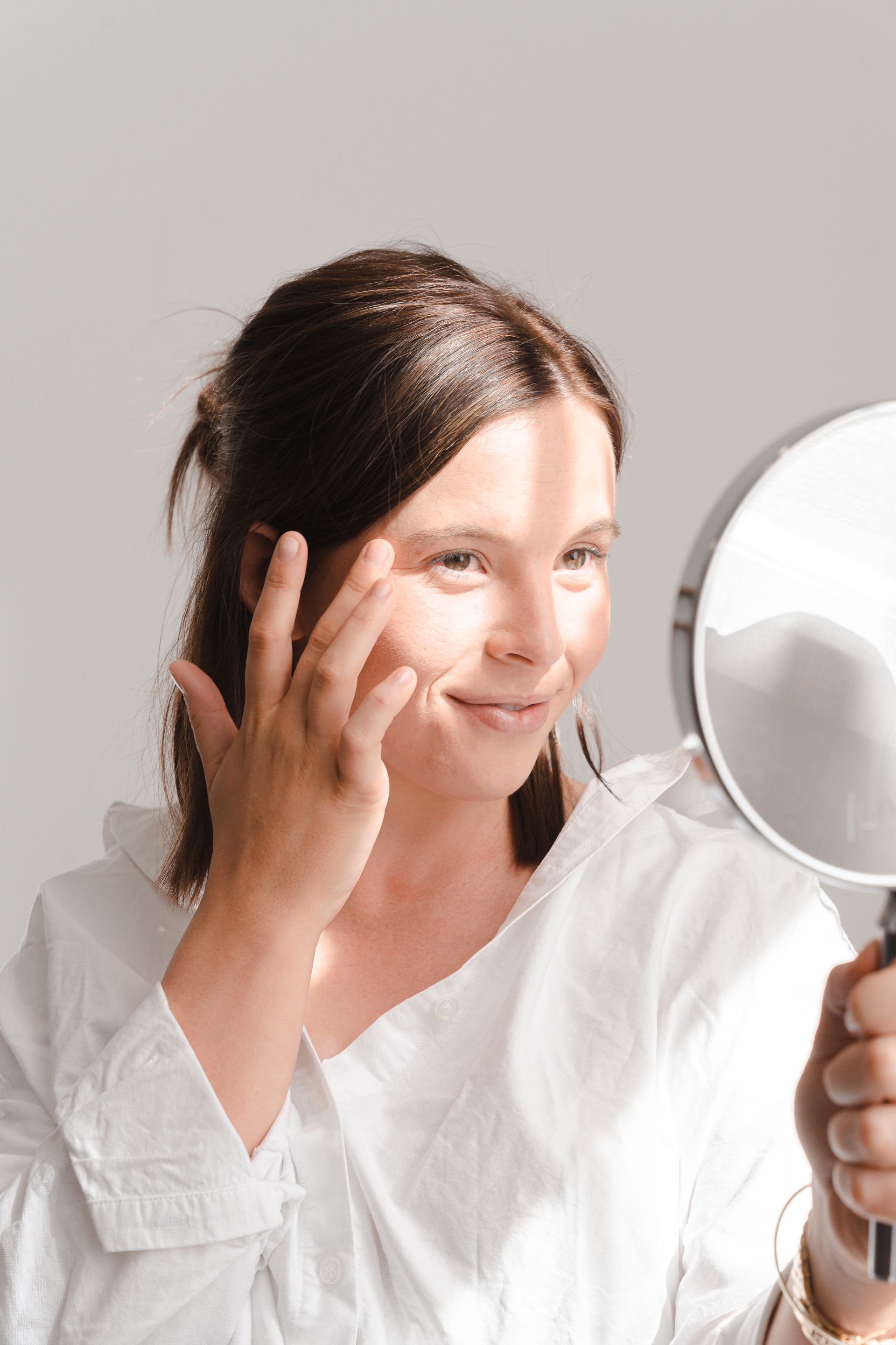 Woman in a white shirt looking into a handheld mirror while touching her face.