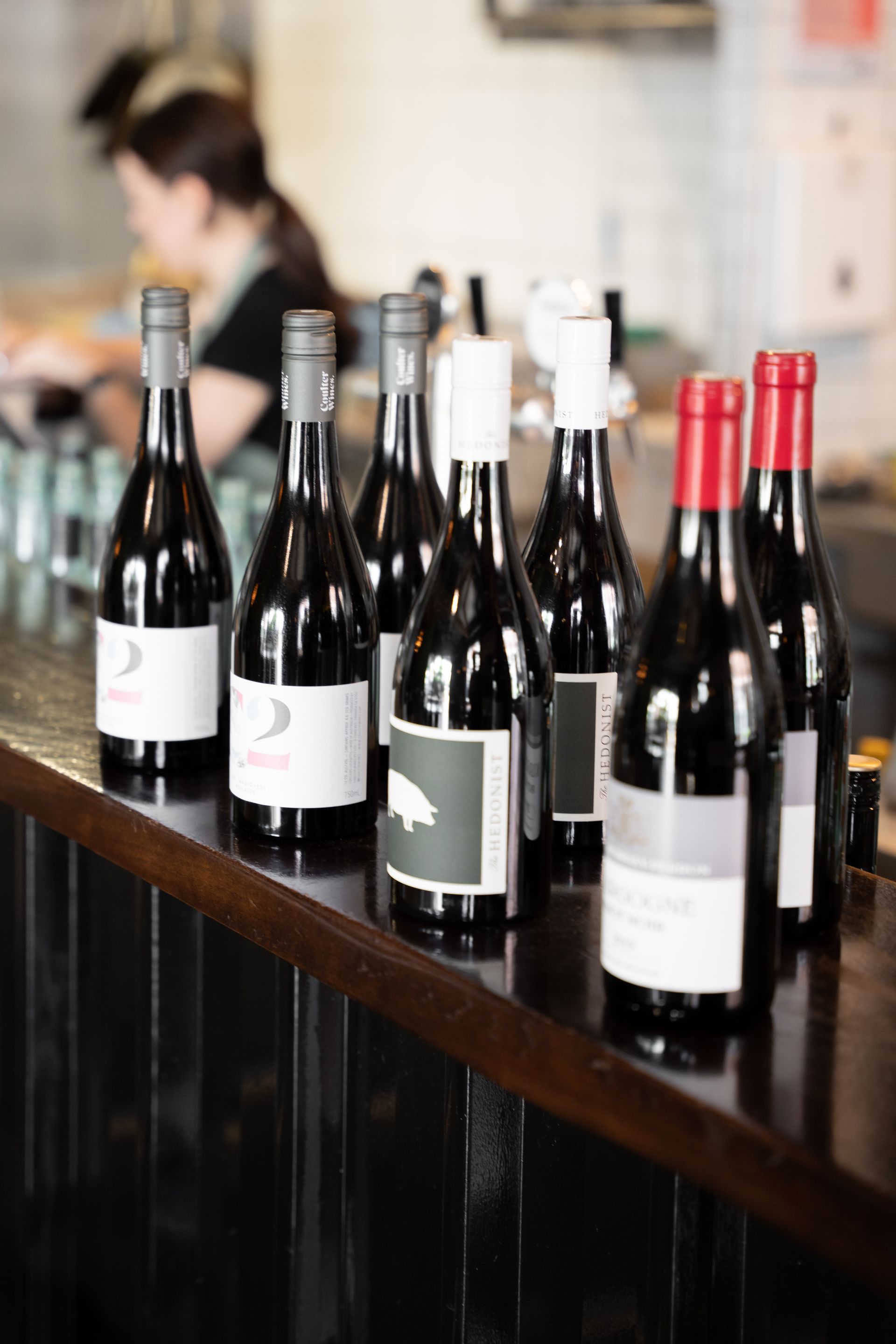 Wine bottles lined up on a counter in a bar or tasting room, with a blurred person in the background.