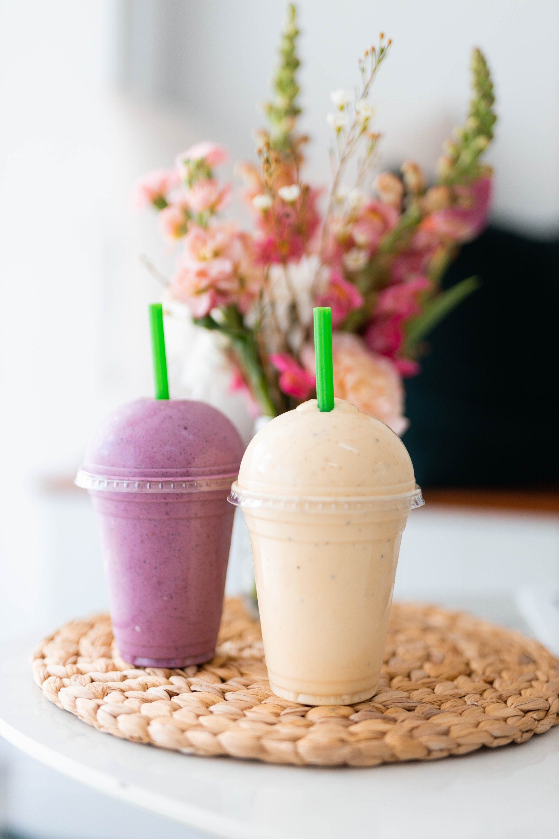 Two pastel smoothies, purple and beige, with green straws on a woven placemat before pink flowers.