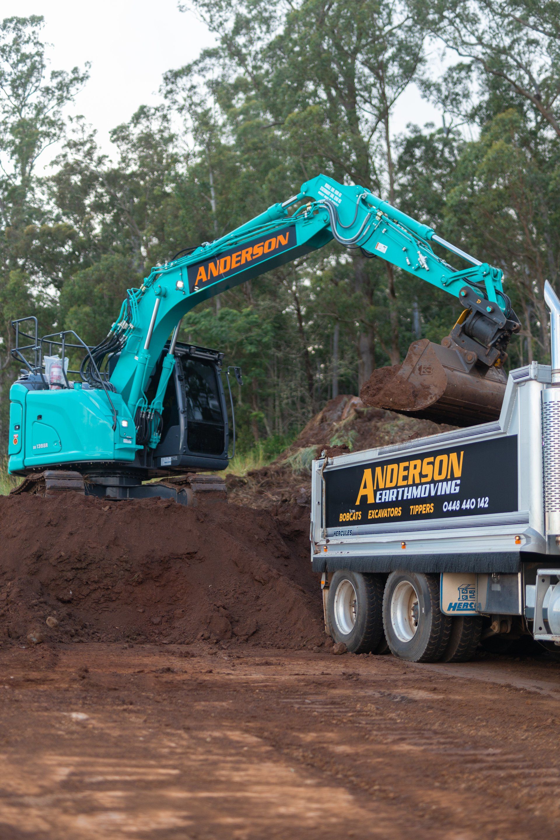 Teal excavator loading dirt into a dump truck at a muddy construction site