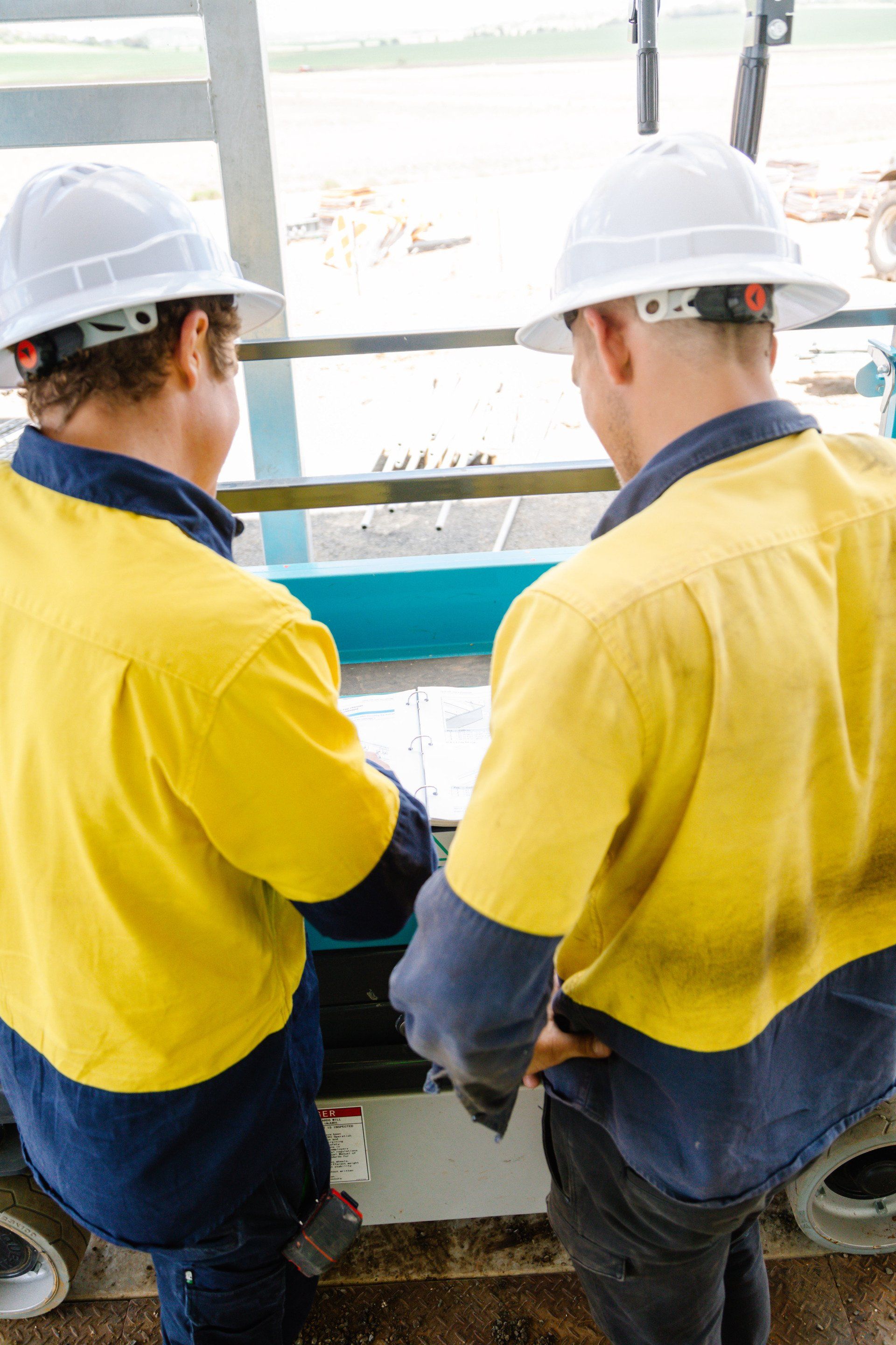 Two workers in yellow and blue uniforms and white hard hats stand at a machine in a factory.