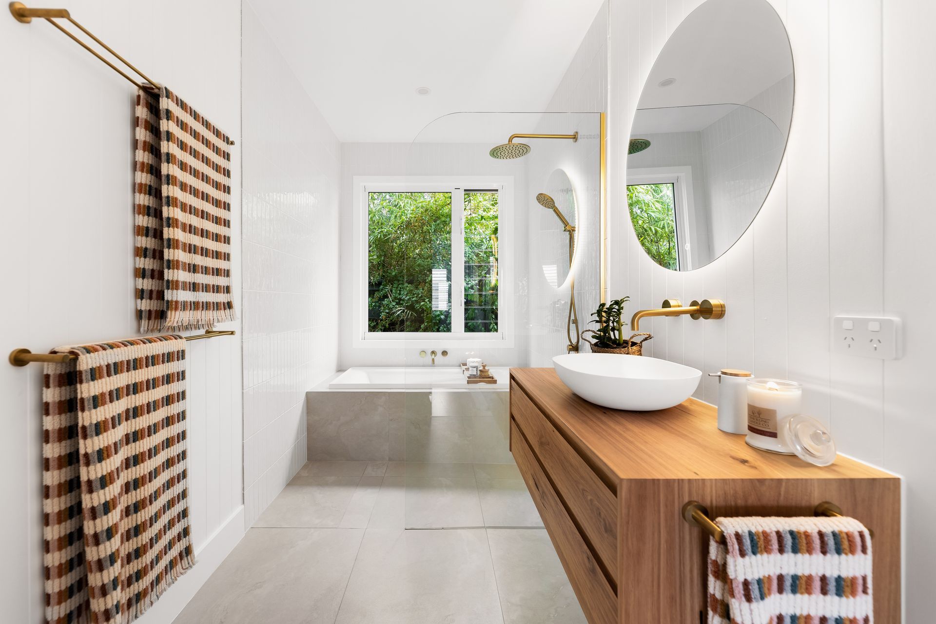 Modern bathroom with wood vanity, round mirror, tub by window, and plaid towels