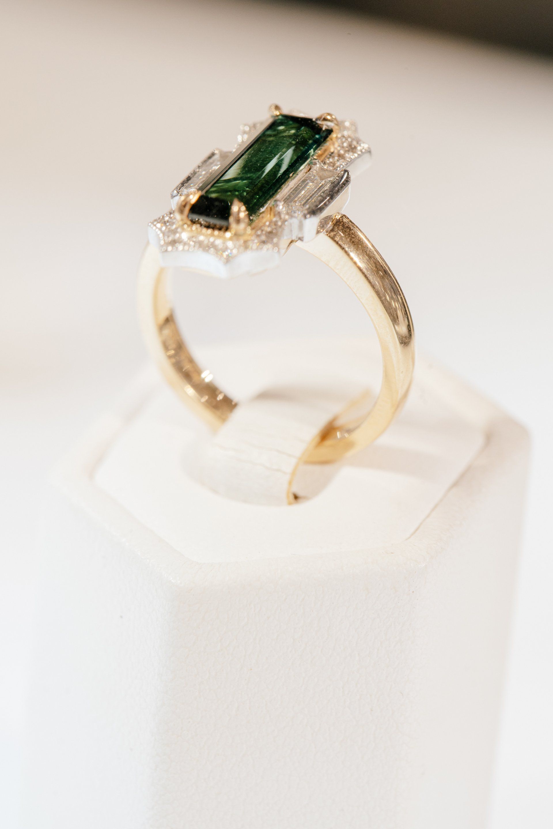 Gold ring with a rectangular green gemstone and diamond accents on a white display stand