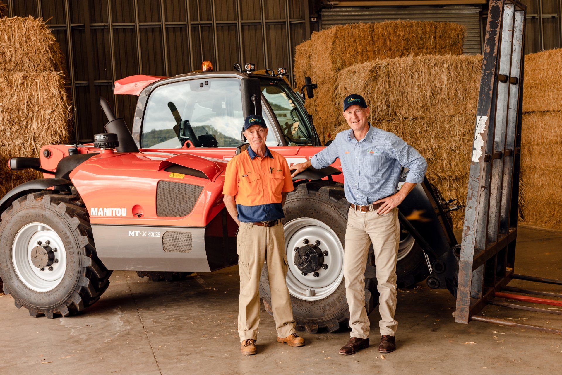 Two people standing beside a red tractor in a barn with stacked hay bales.