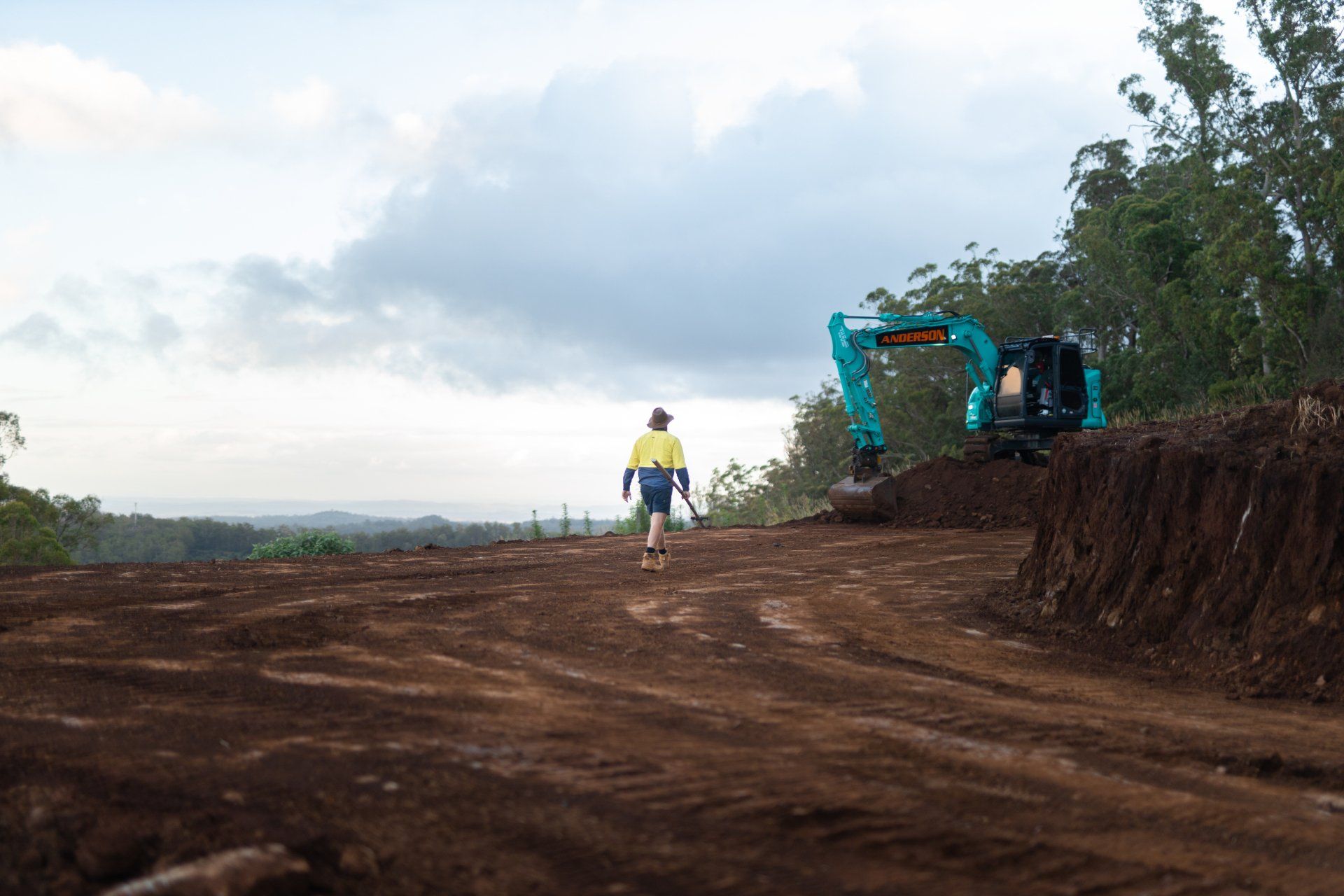 Worker and blue excavator on a dirt road by a hillside under a cloudy sky