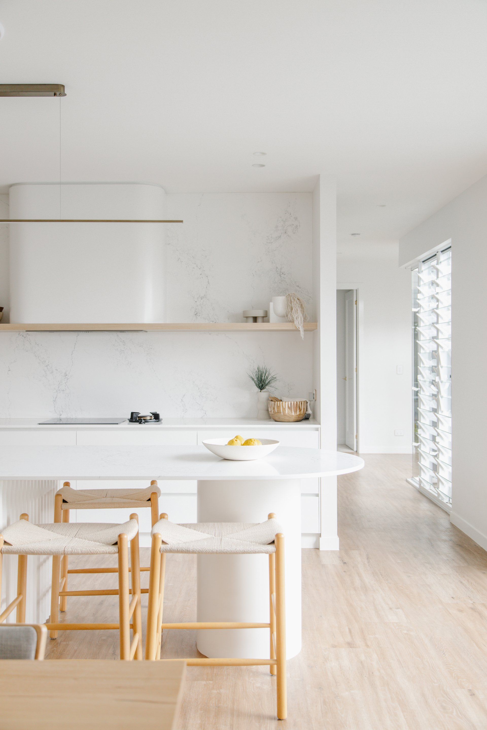 Bright minimalist kitchen with white island, wooden chairs, open shelves, and soft natural light
