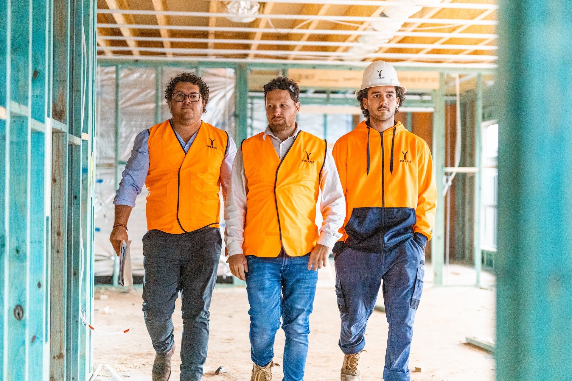 Three construction workers in orange safety vests walking through a building under construction