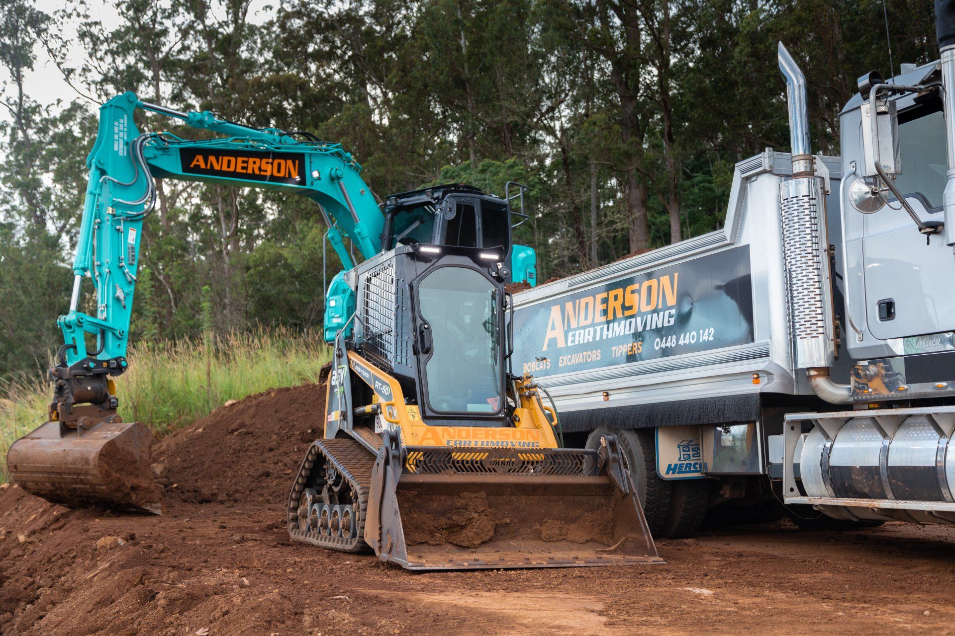 Blue excavator and white dump truck on a dirt road in a wooded area.