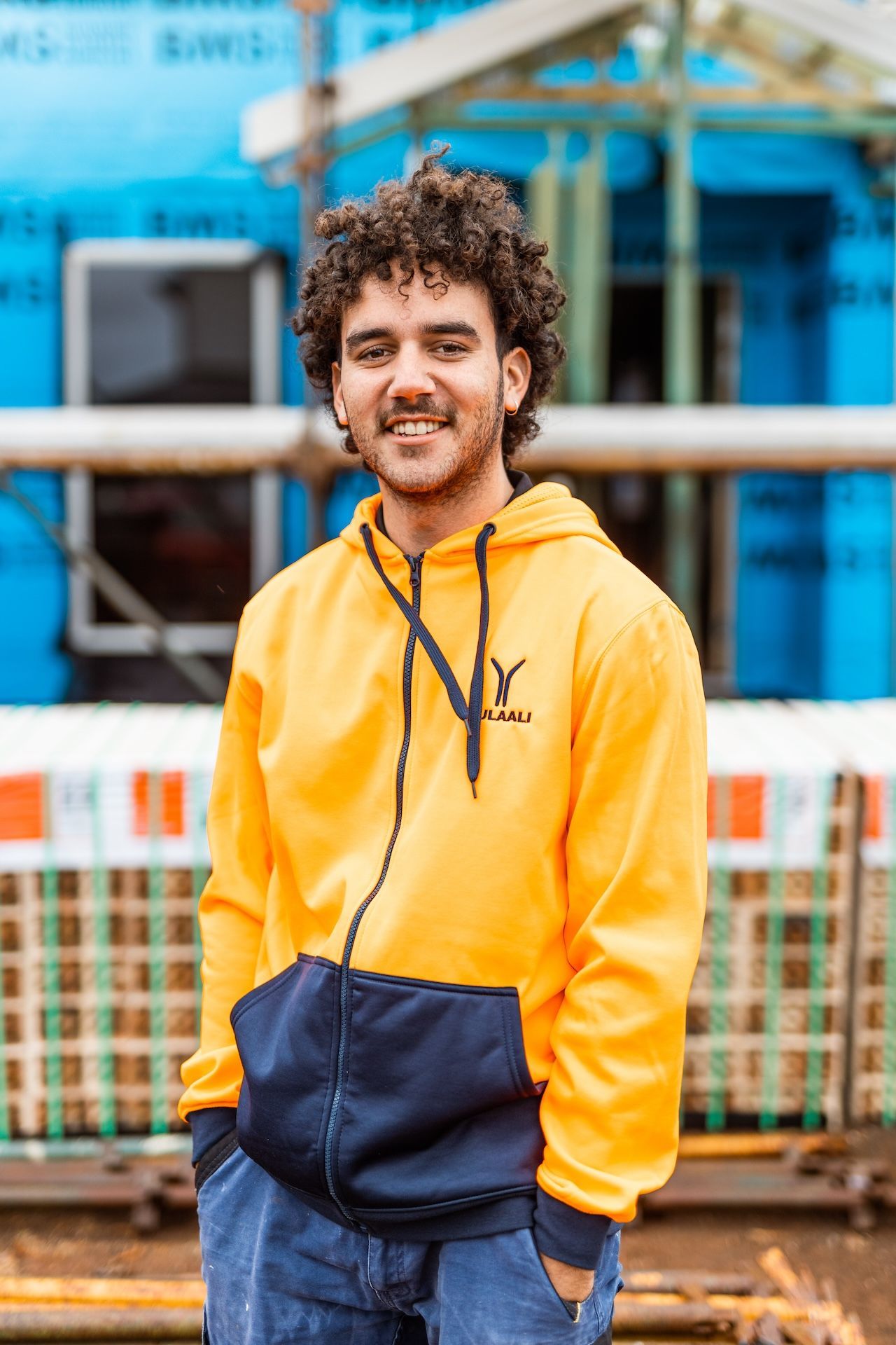 Man in a yellow hoodie standing outdoors in front of blue industrial equipment