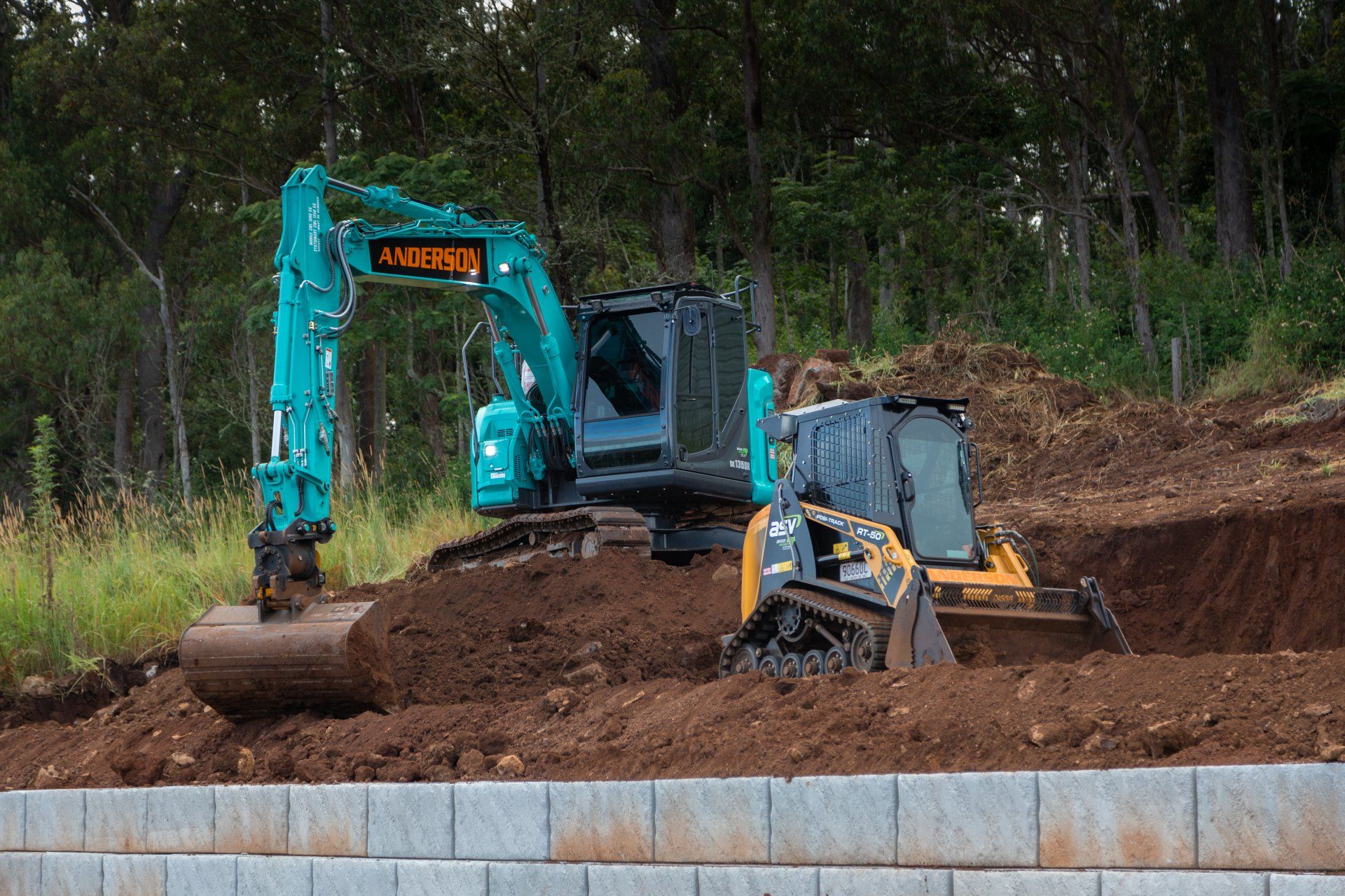 Turquoise excavator digging dirt beside a retaining wall in a wooded construction site