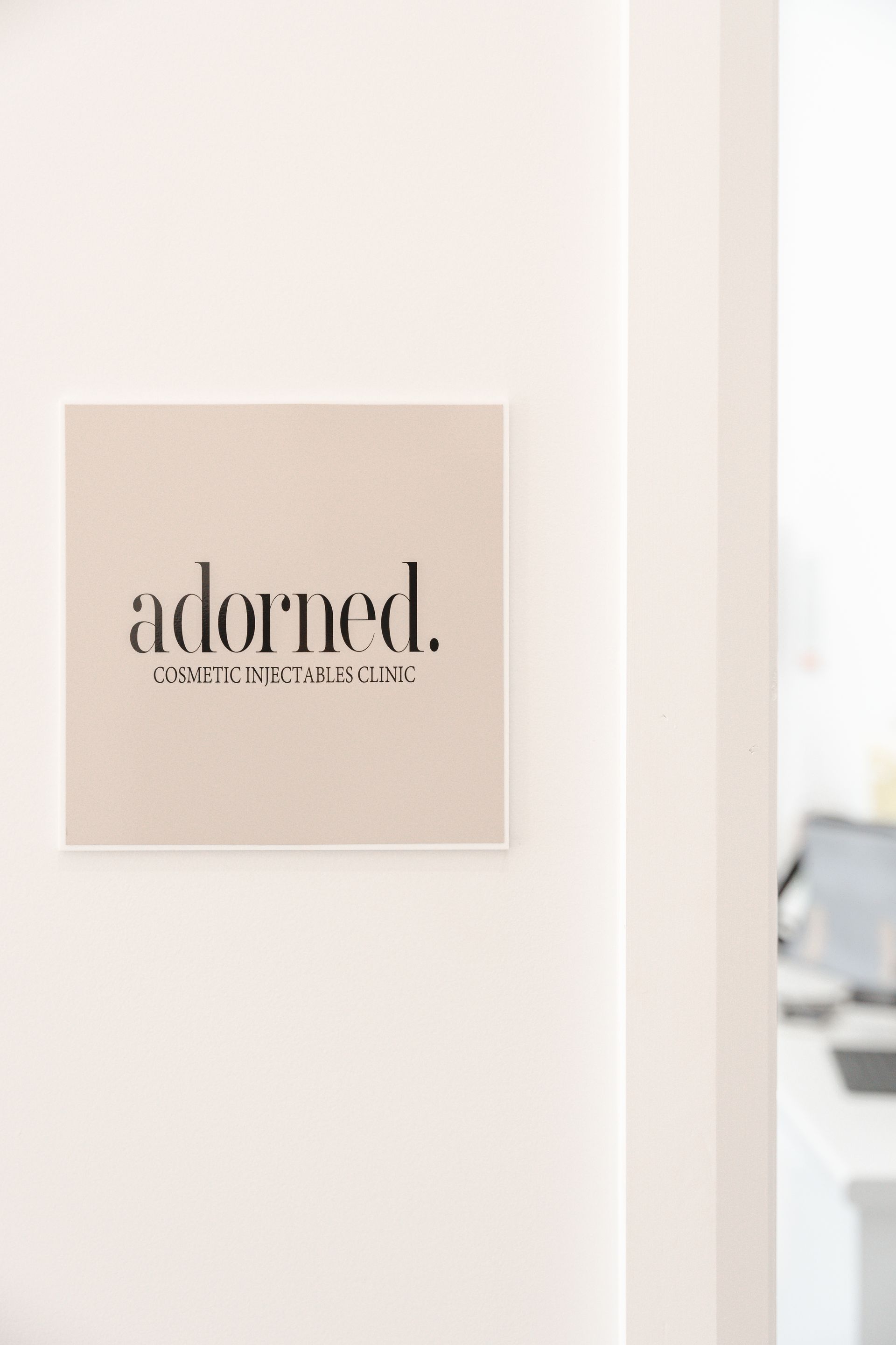 Small beige wall sign reading “adorned.” in a bright white interior room
