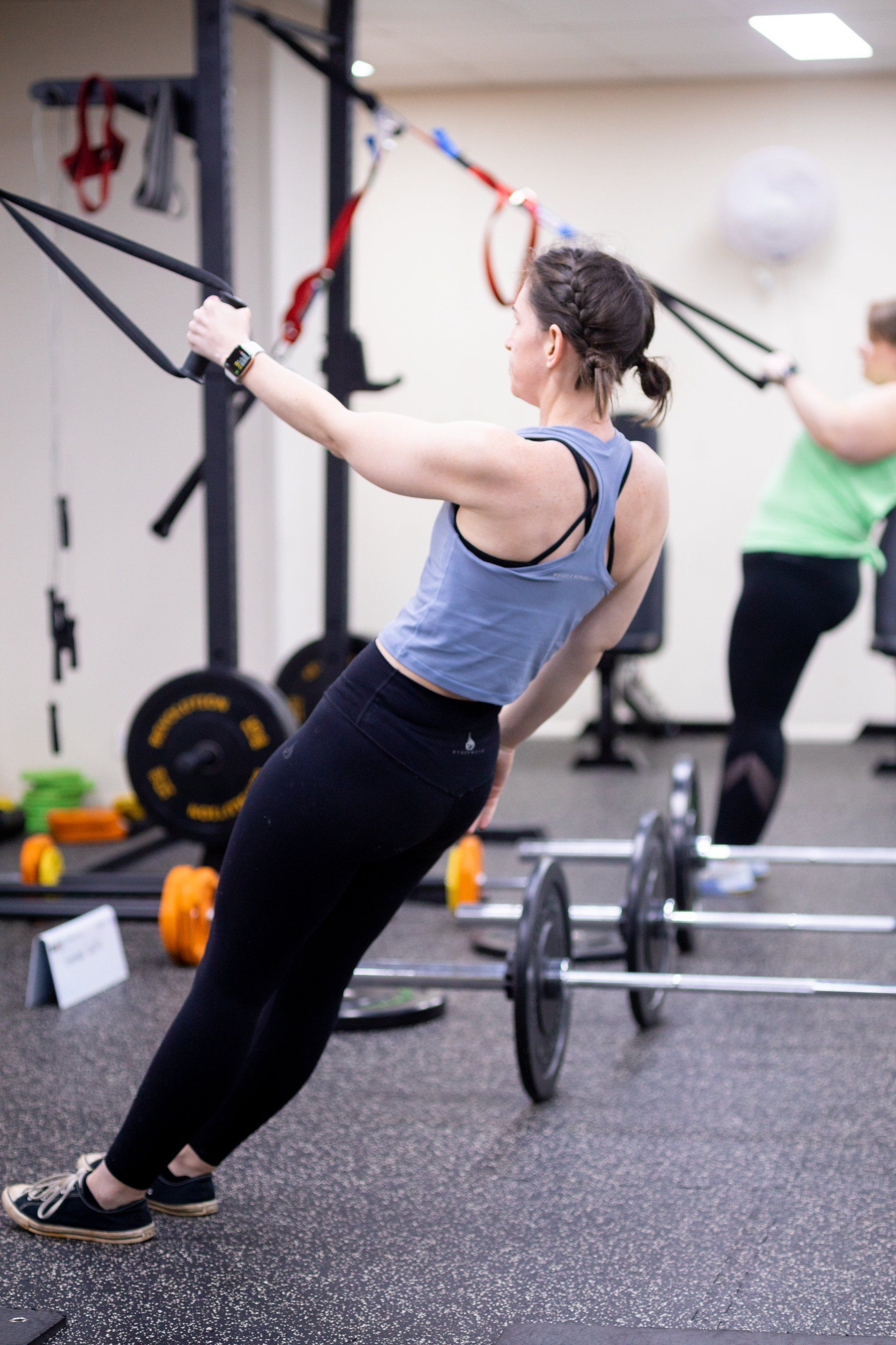 Woman doing TRX rows in a gym, leaning back and holding suspension straps near barbells.