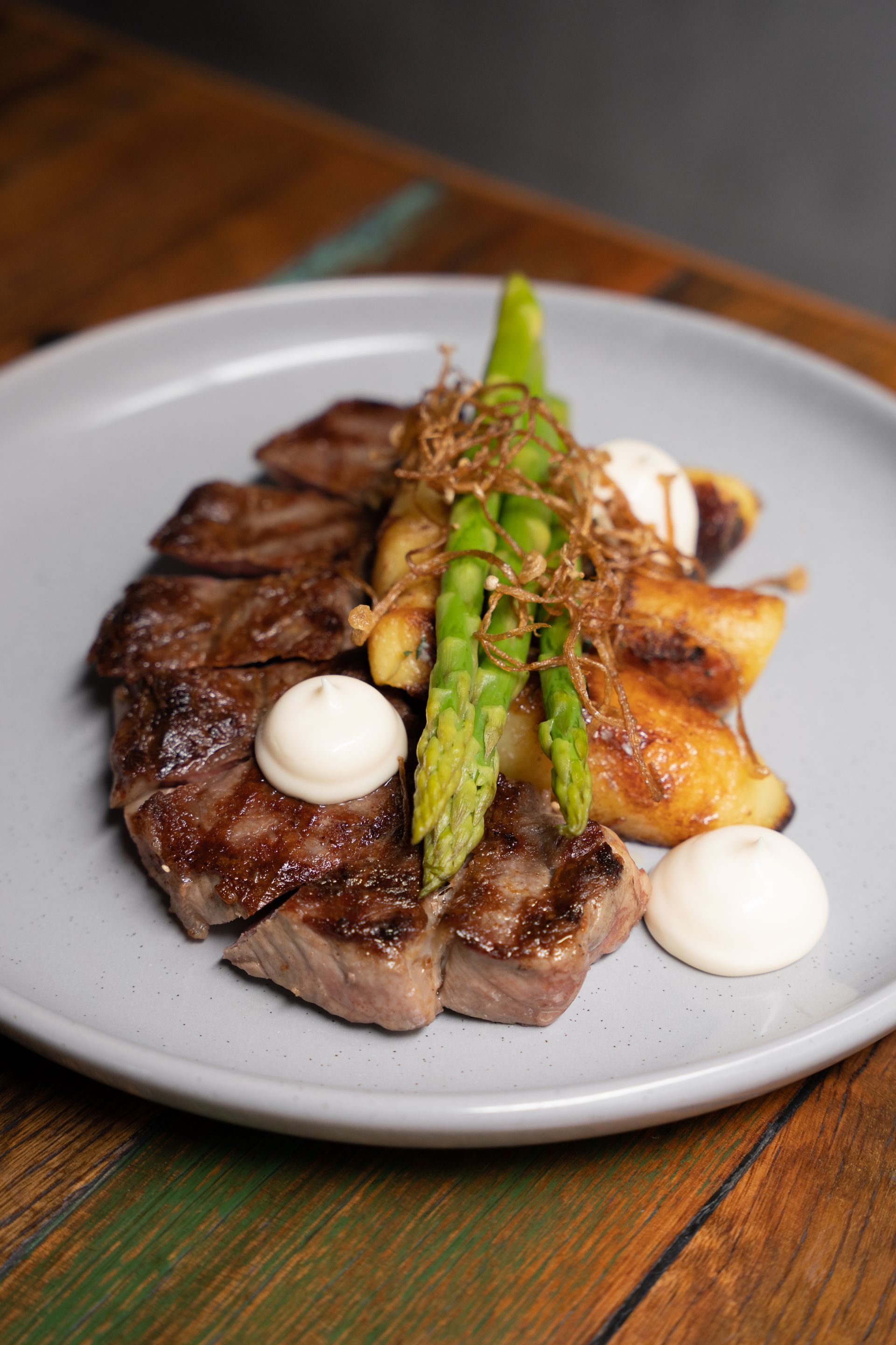 Grilled steak with asparagus and dollops of sauce on a white plate on a wooden table
