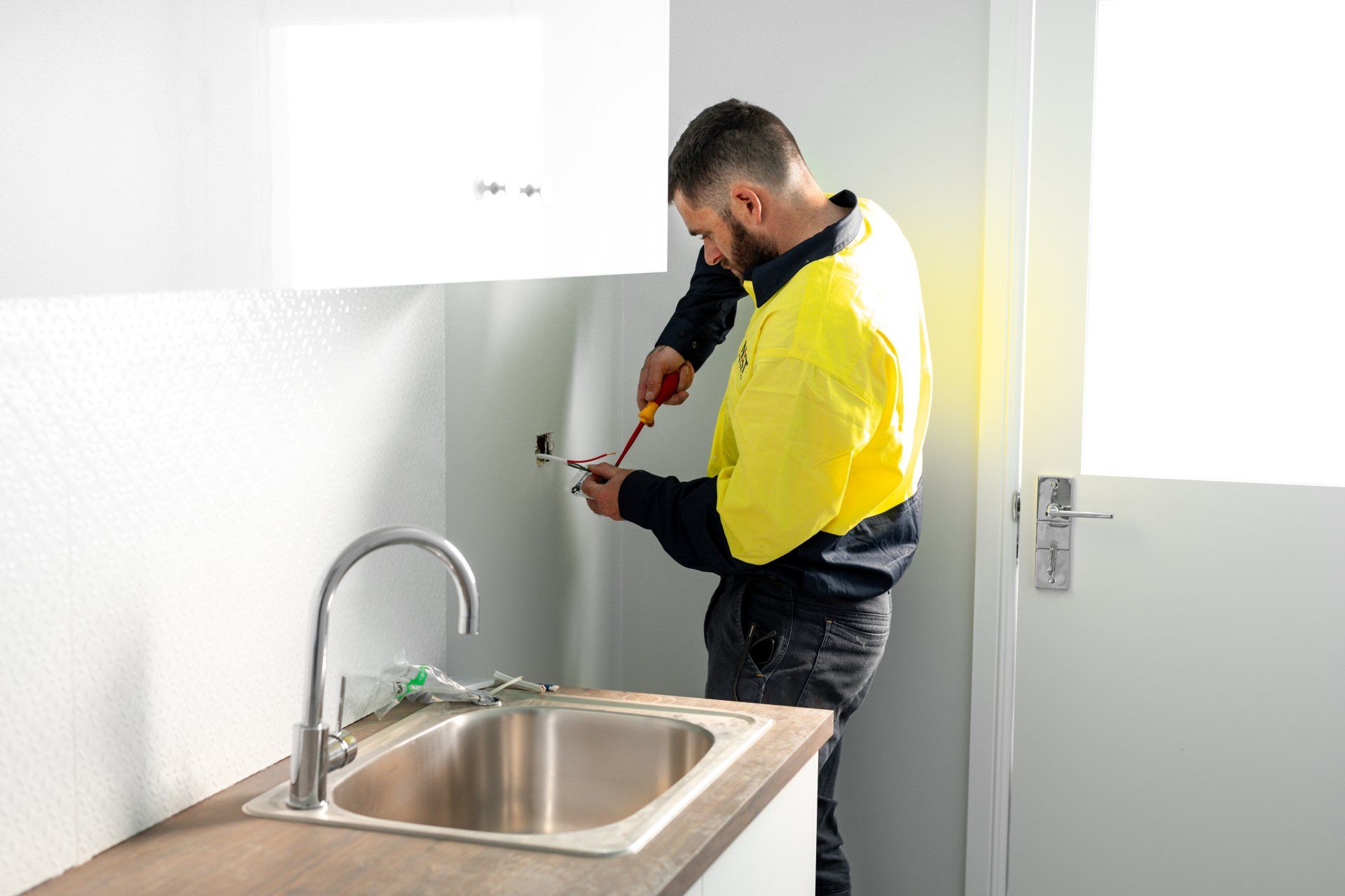 Worker in a yellow jacket repairing a kitchen sink cabinet with a drill
