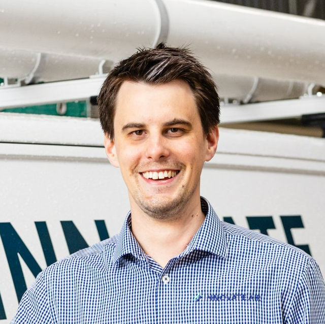 Smiling man in blue checkered shirt standing indoors with a company sign behind him