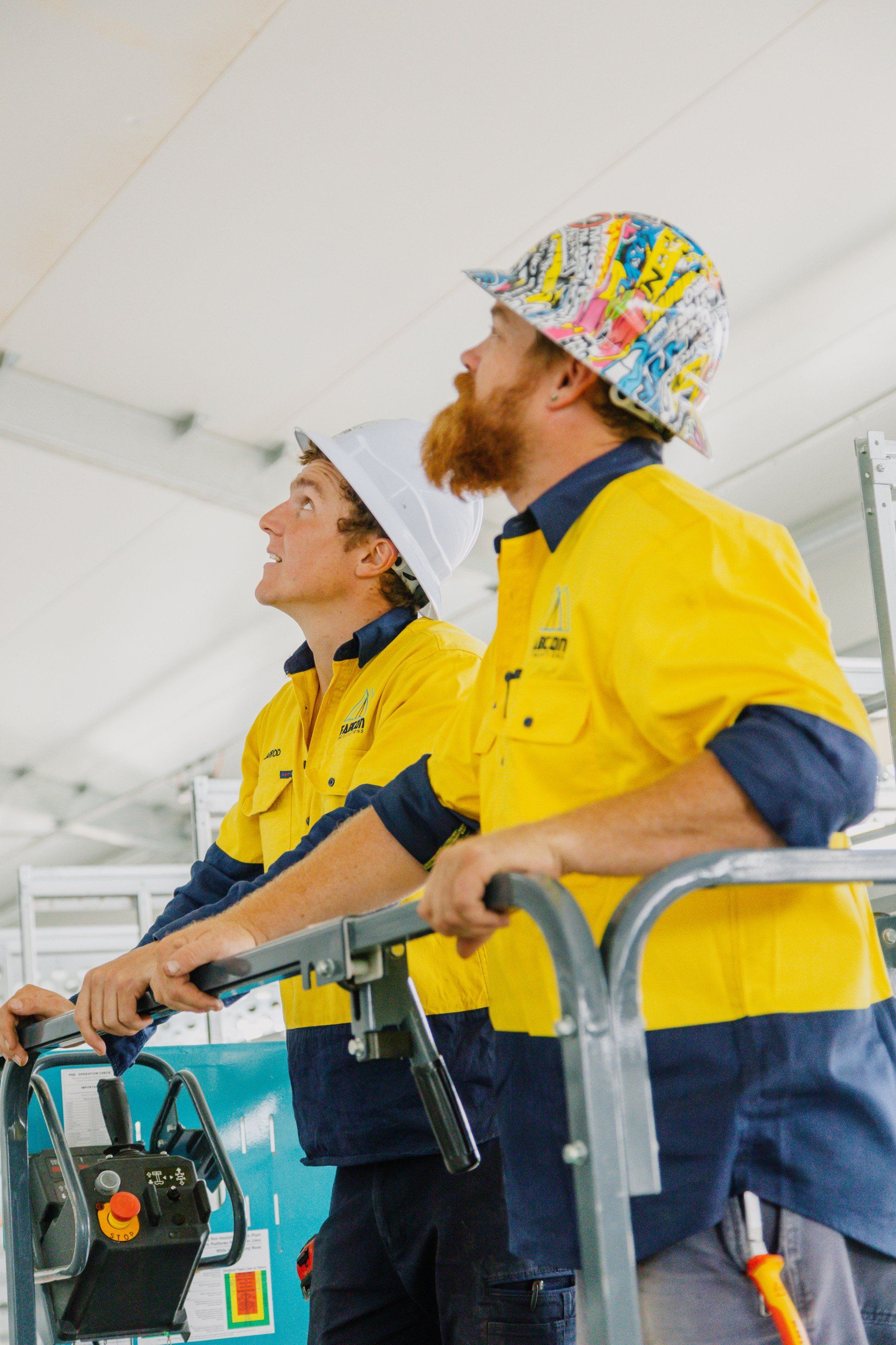 Two workers in yellow safety jackets and hard hats inspecting machinery on a worksite.