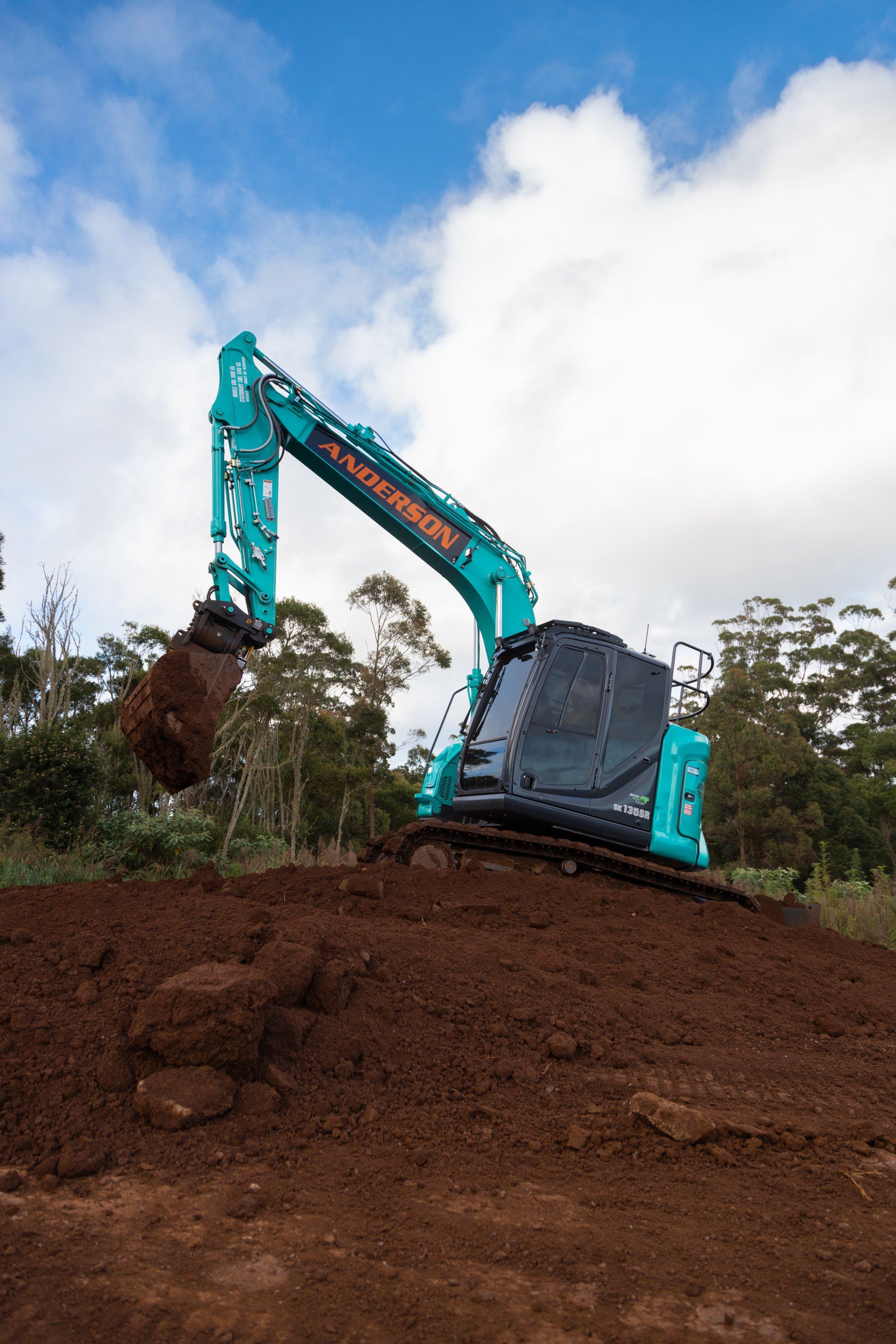 Turquoise excavator digging on a dirt mound under a cloudy sky