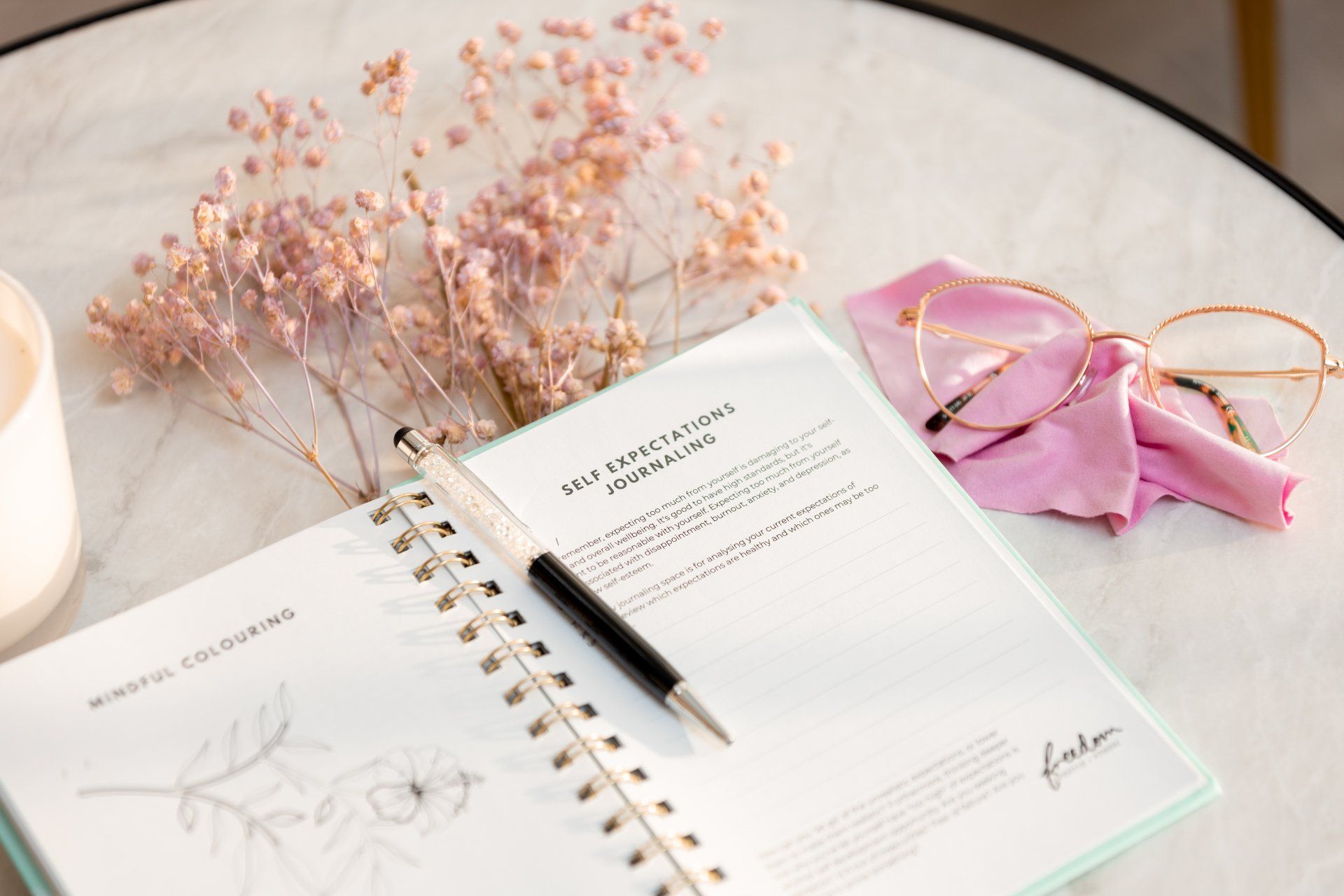 Open spiral planner with pen, pink glasses, and dried flowers on a white desk