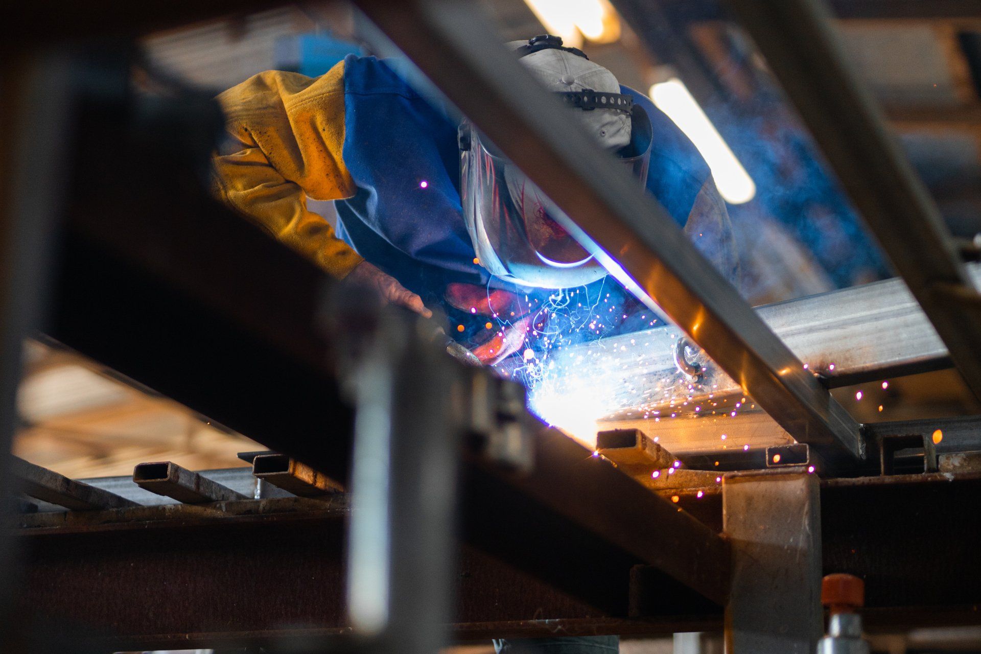 Person welding metal under a workbench, with bright blue sparks and orange light in a workshop