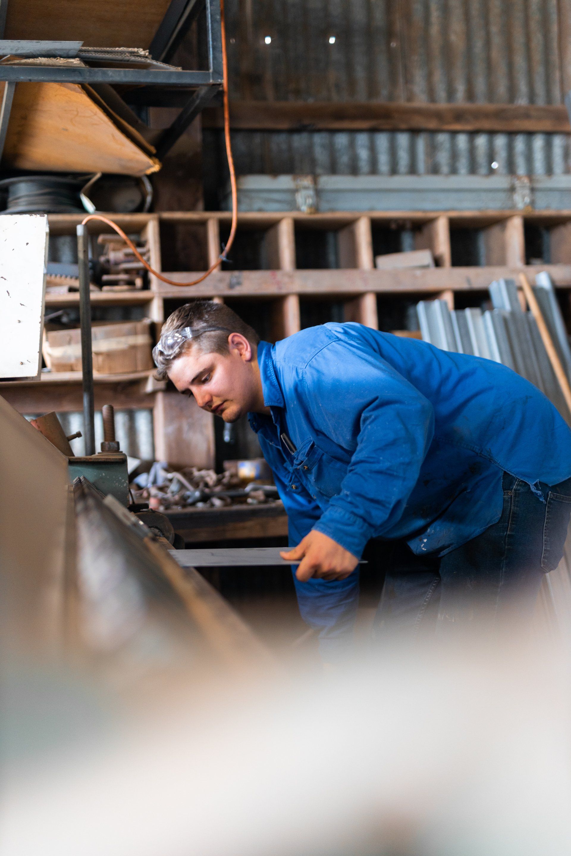 Man in a blue shirt leaning over a workbench in a workshop with tools and shelves behind him