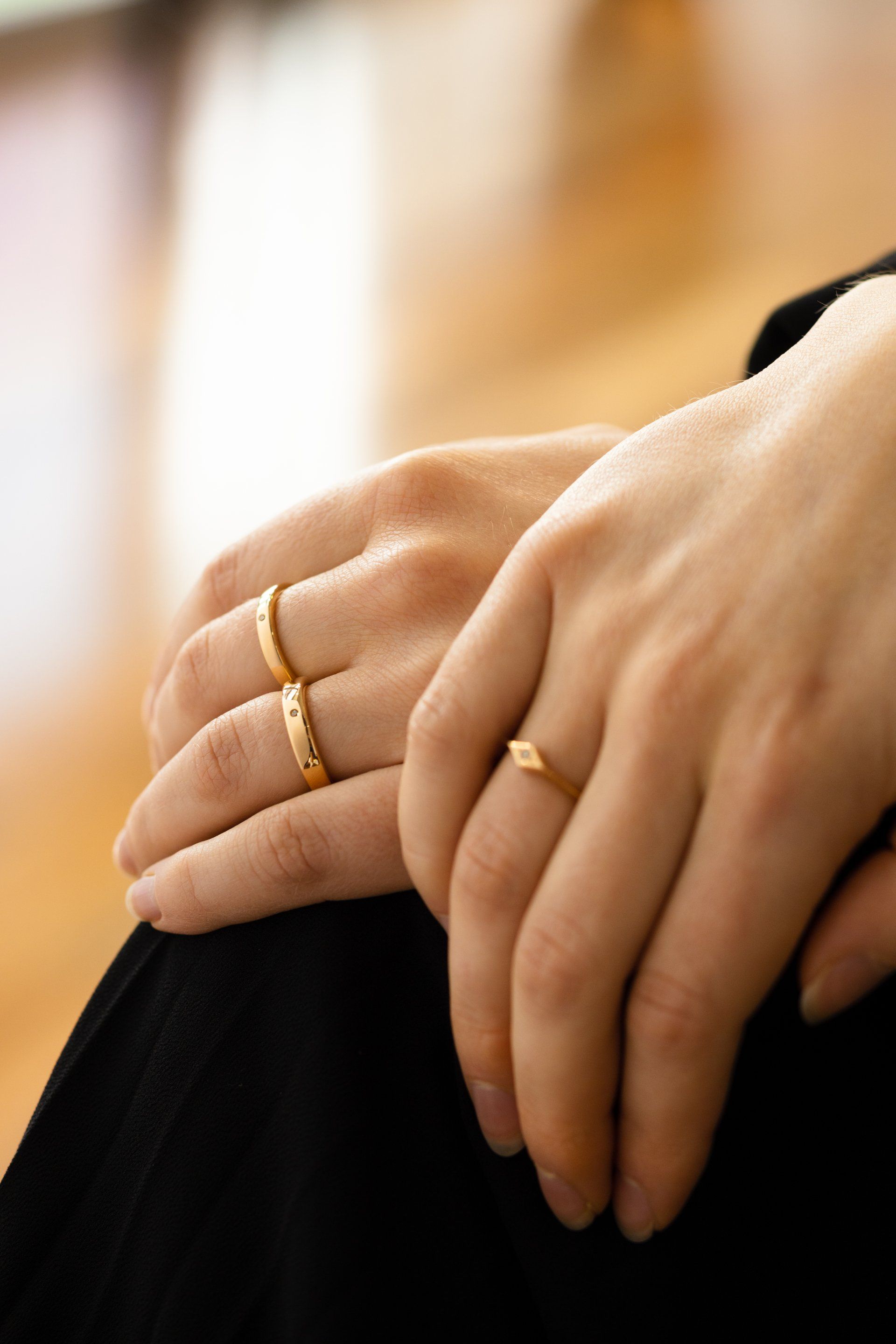 Hands resting together, wearing gold rings, against a soft blurred background.