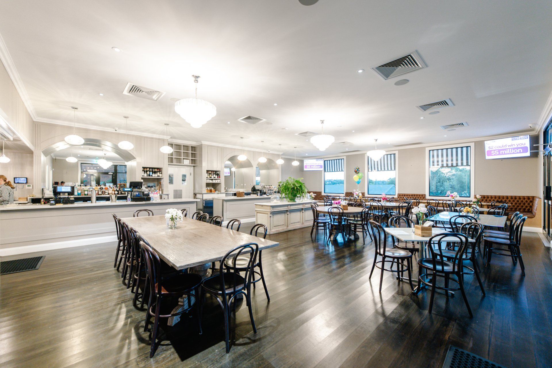 Bright community dining hall with long tables, black chairs, and a serving counter under ceiling lights