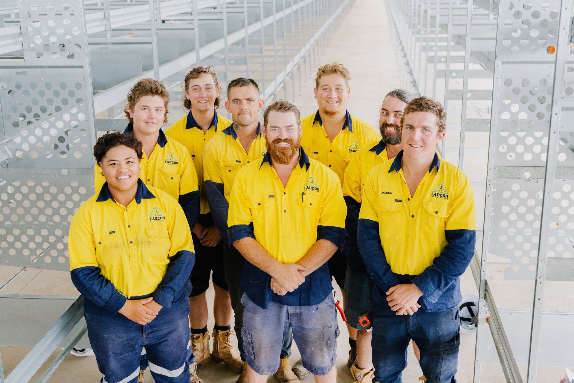 Seven workers in yellow shirts standing on a metal walkway in a bright industrial facility