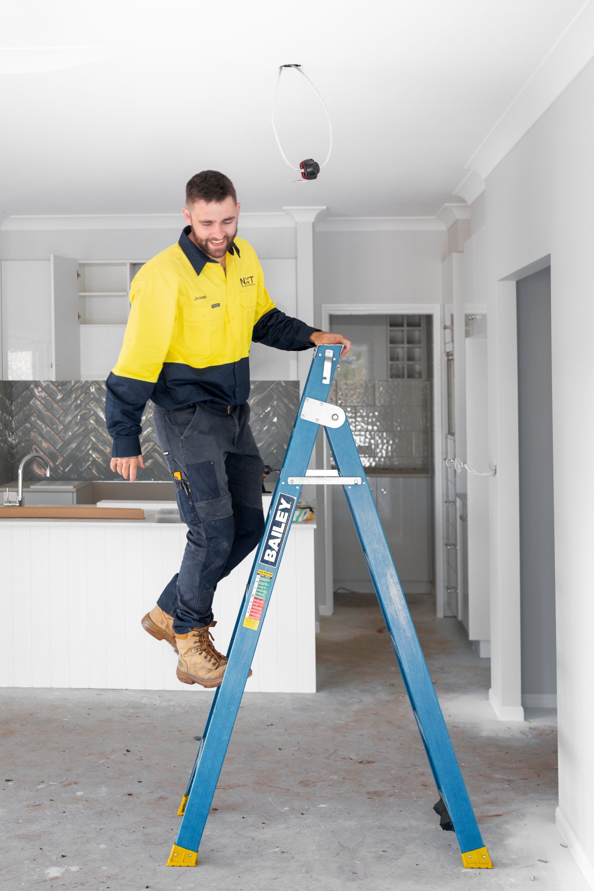 Man in yellow jacket standing on a ladder in a kitchen under construction, holding a tool in hand