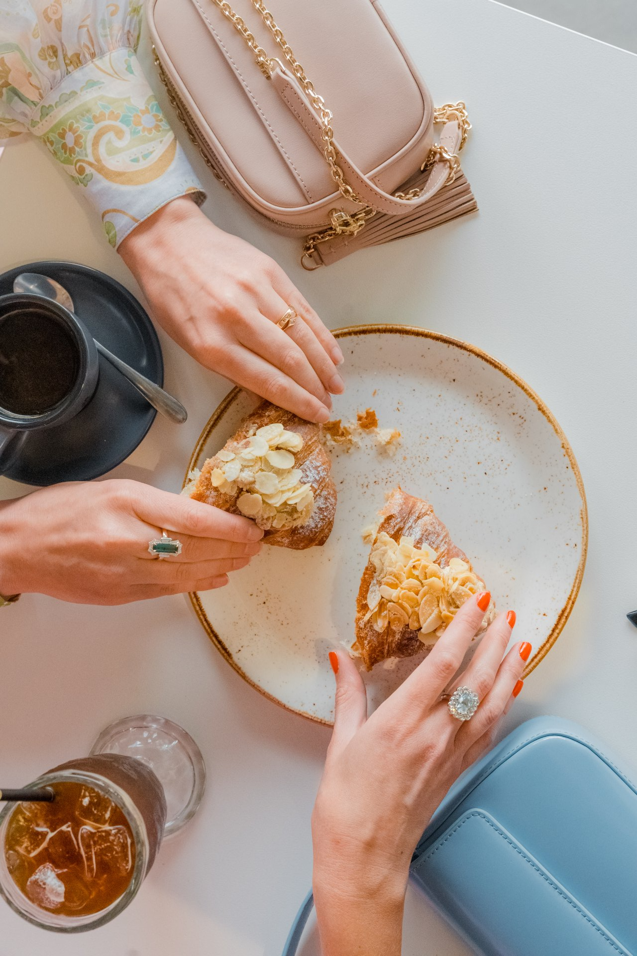 Two hands sharing pastries at a table with tea, coffee, and a pink handbag
