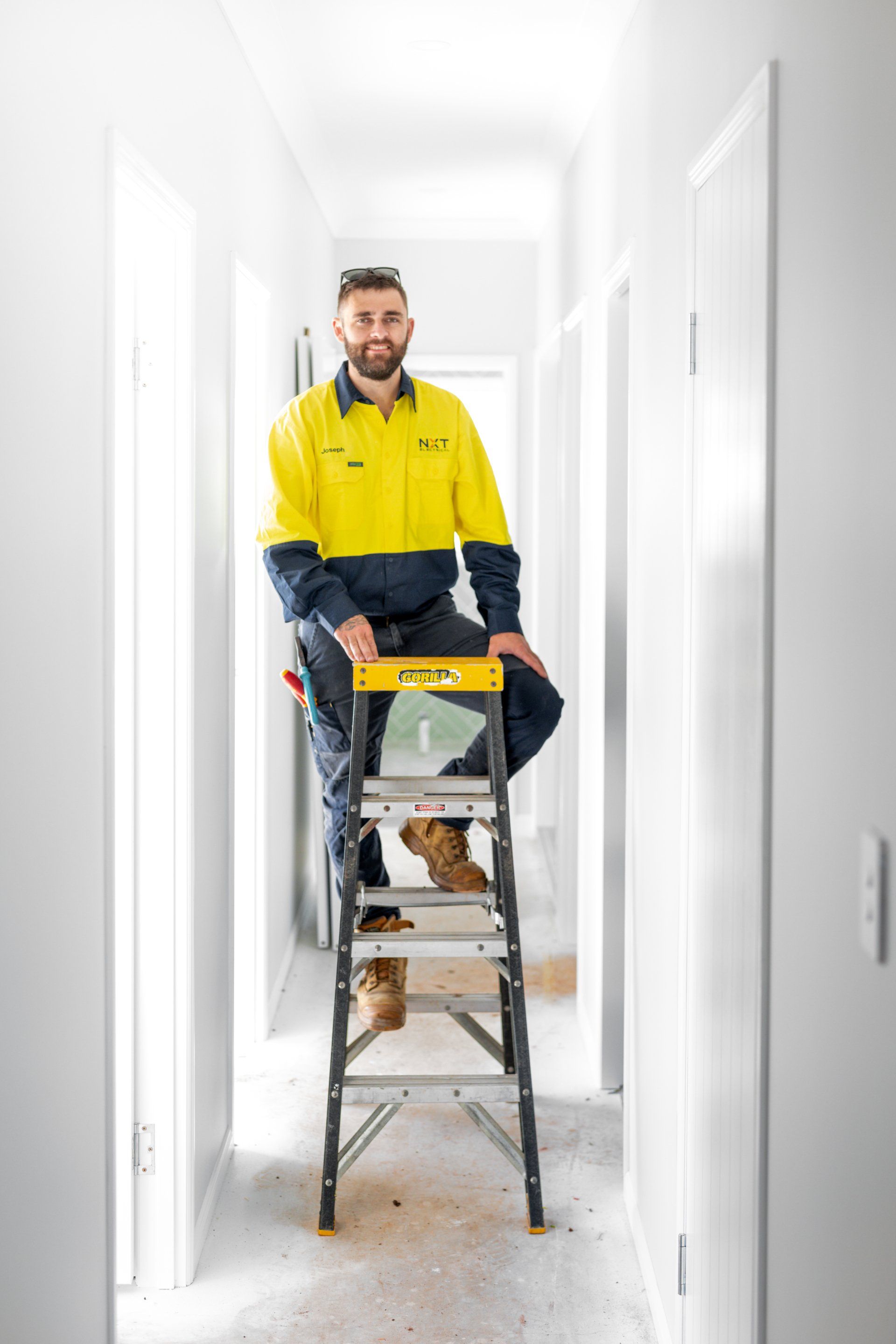 Worker in yellow jacket standing on a ladder in a white hallway, holding a tool on top of the ladder