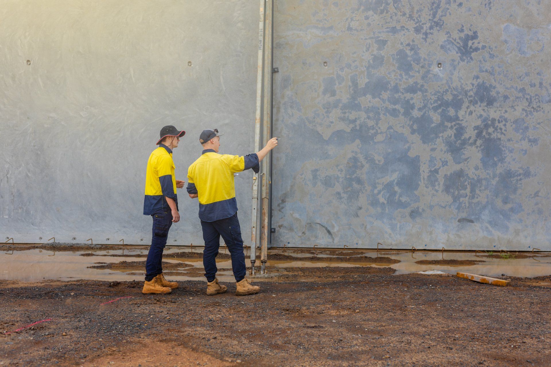Two workers in yellow shirts inspect a large gray wall, one pointing at a vertical seam.